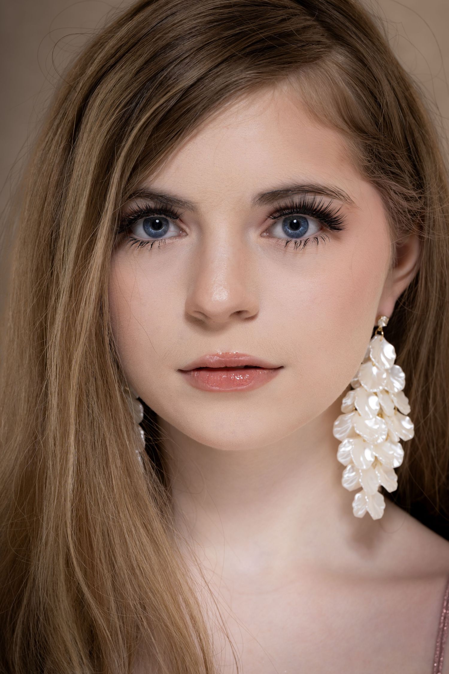 Young woman with blue eyes and long blonde hair wearing elegant white floral drop earrings, soft makeup, neutral background.
