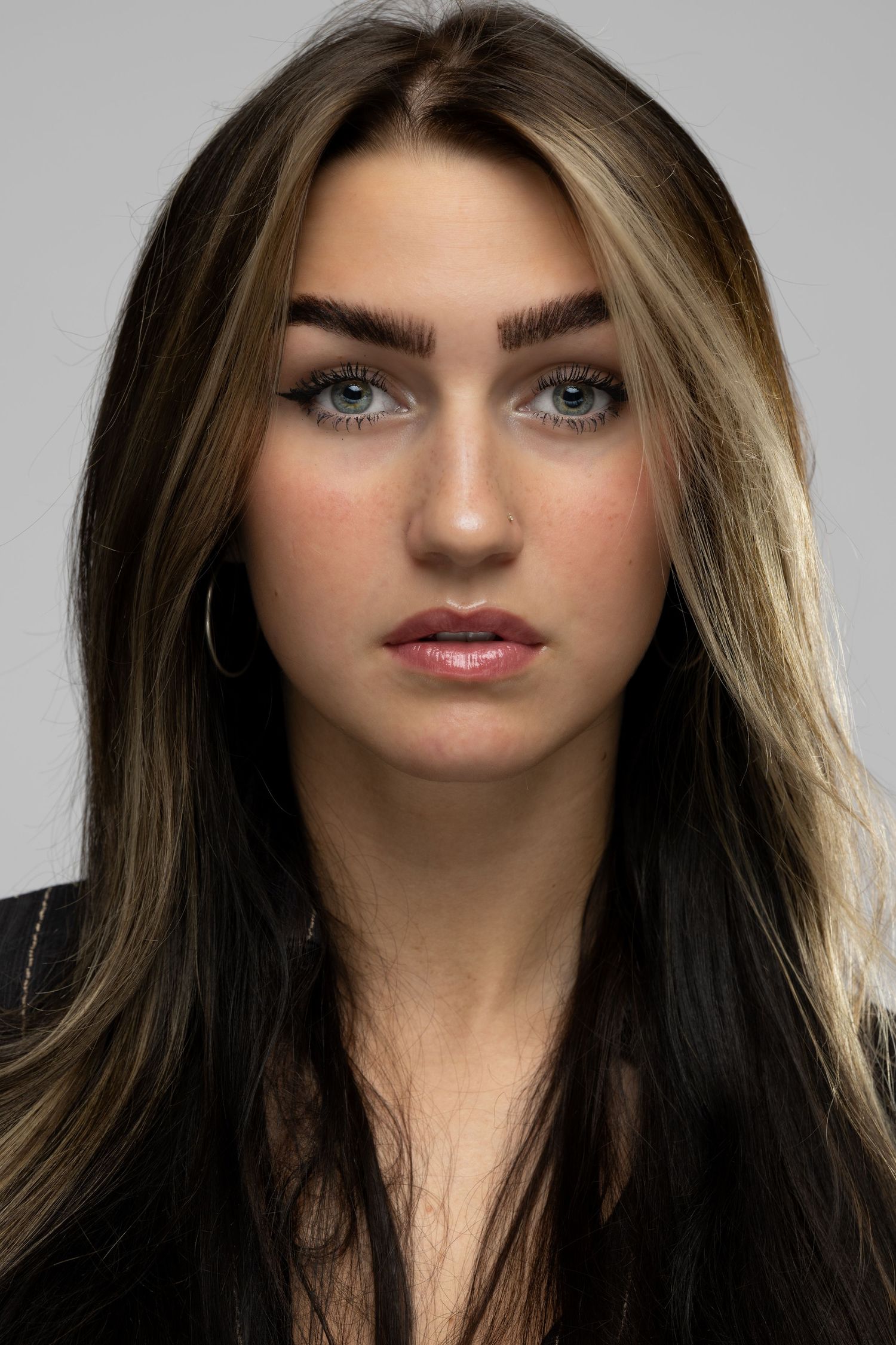 Young woman with long dark hair and blonde highlights, blue-green eyes, hoop earrings, wearing black top against gray background.