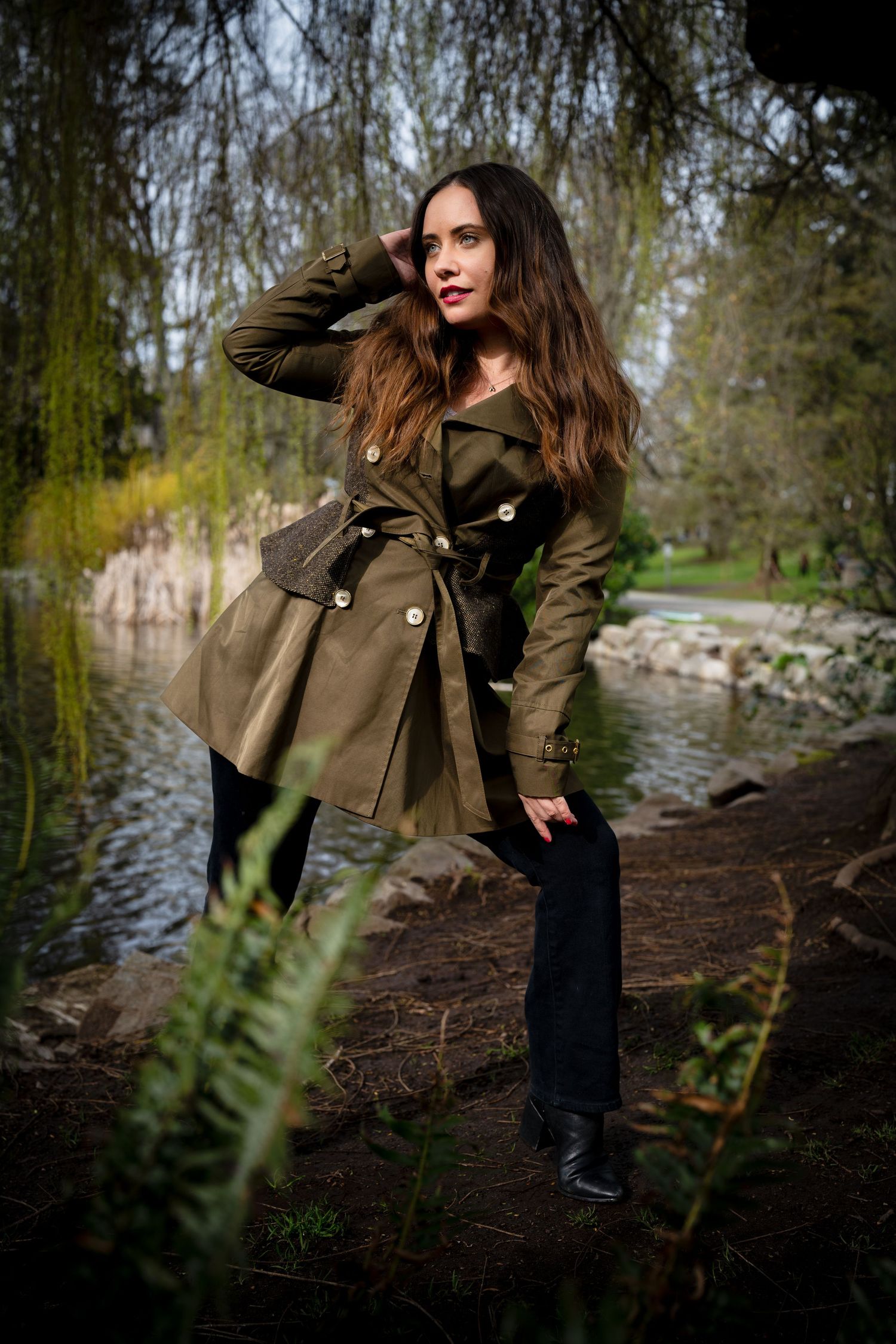Person in olive coat poses dramatically by a river on an overcast day.