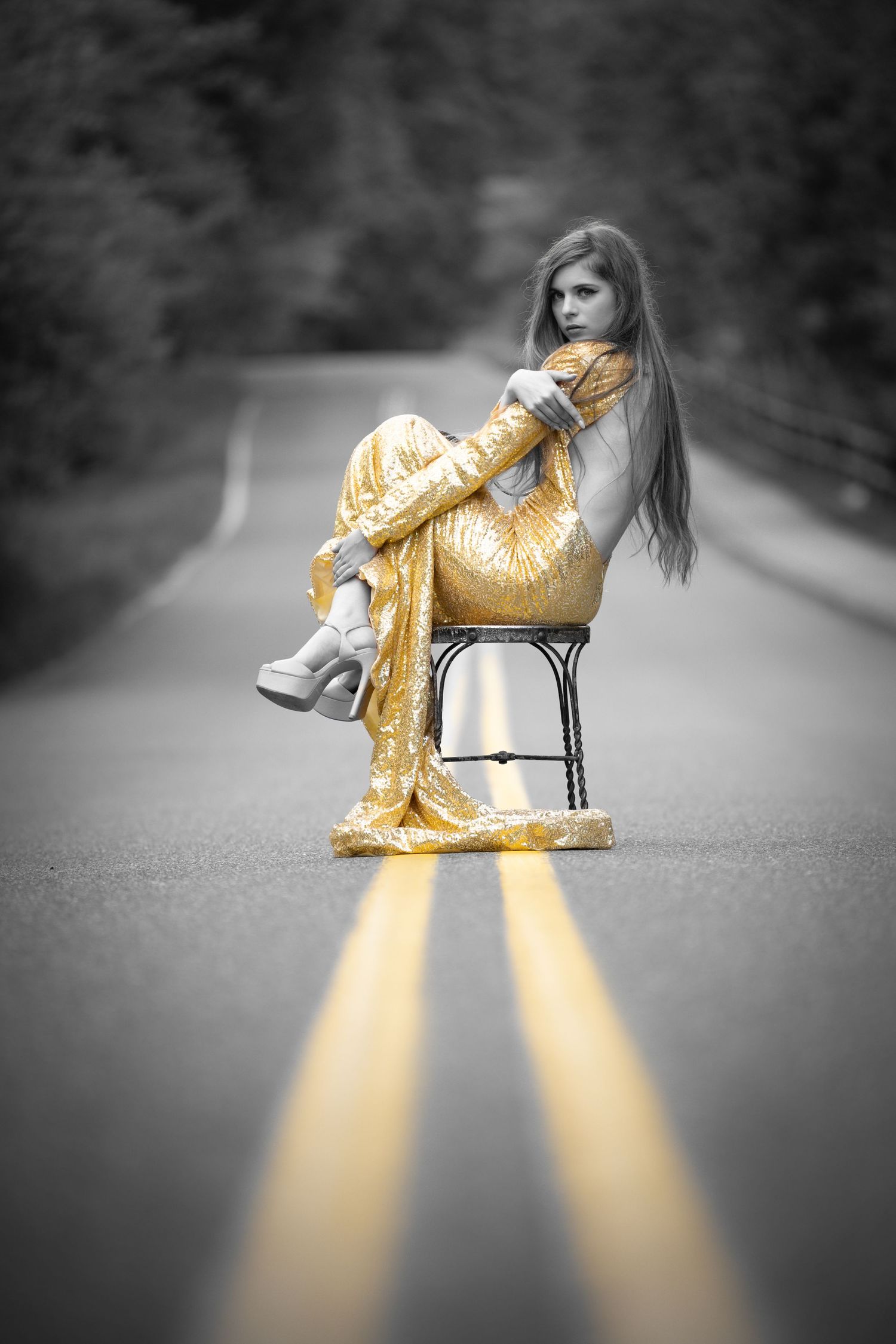 A young woman in a golden dress sits on a chair in the middle of a road, with yellow lines beneath her.