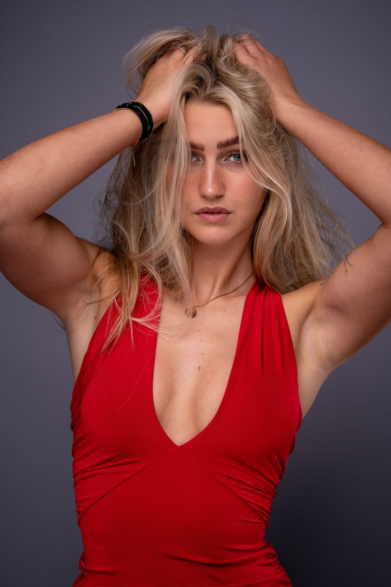 Blonde woman in red deep-v dress poses with hands in hair against gray background.