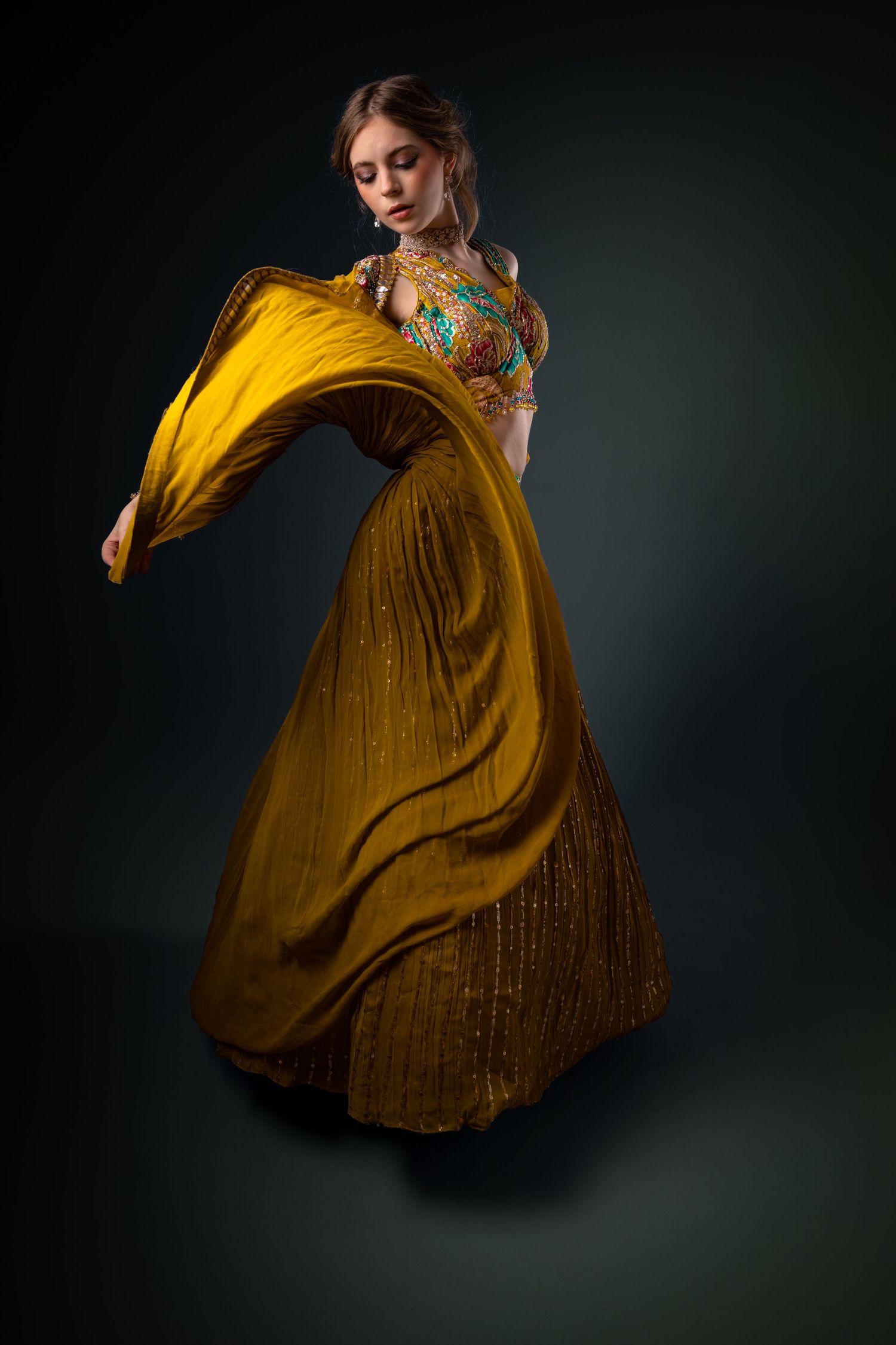 Woman in golden yellow lehenga with colorful embroidered blouse, twirling gracefully against dark background.