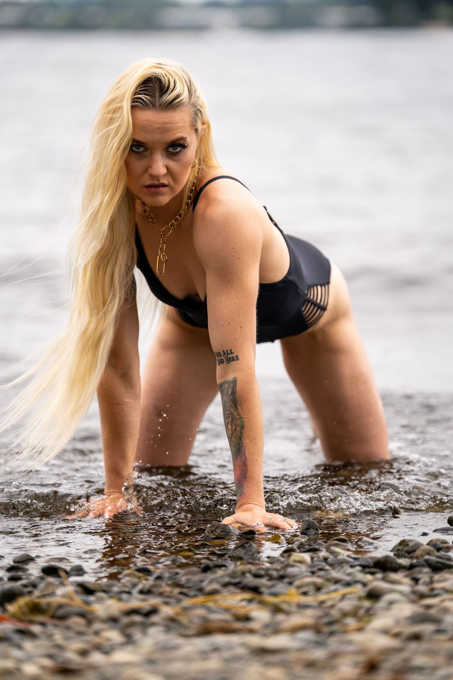 Model in black swimwear poses in shallow water on rocky beach.