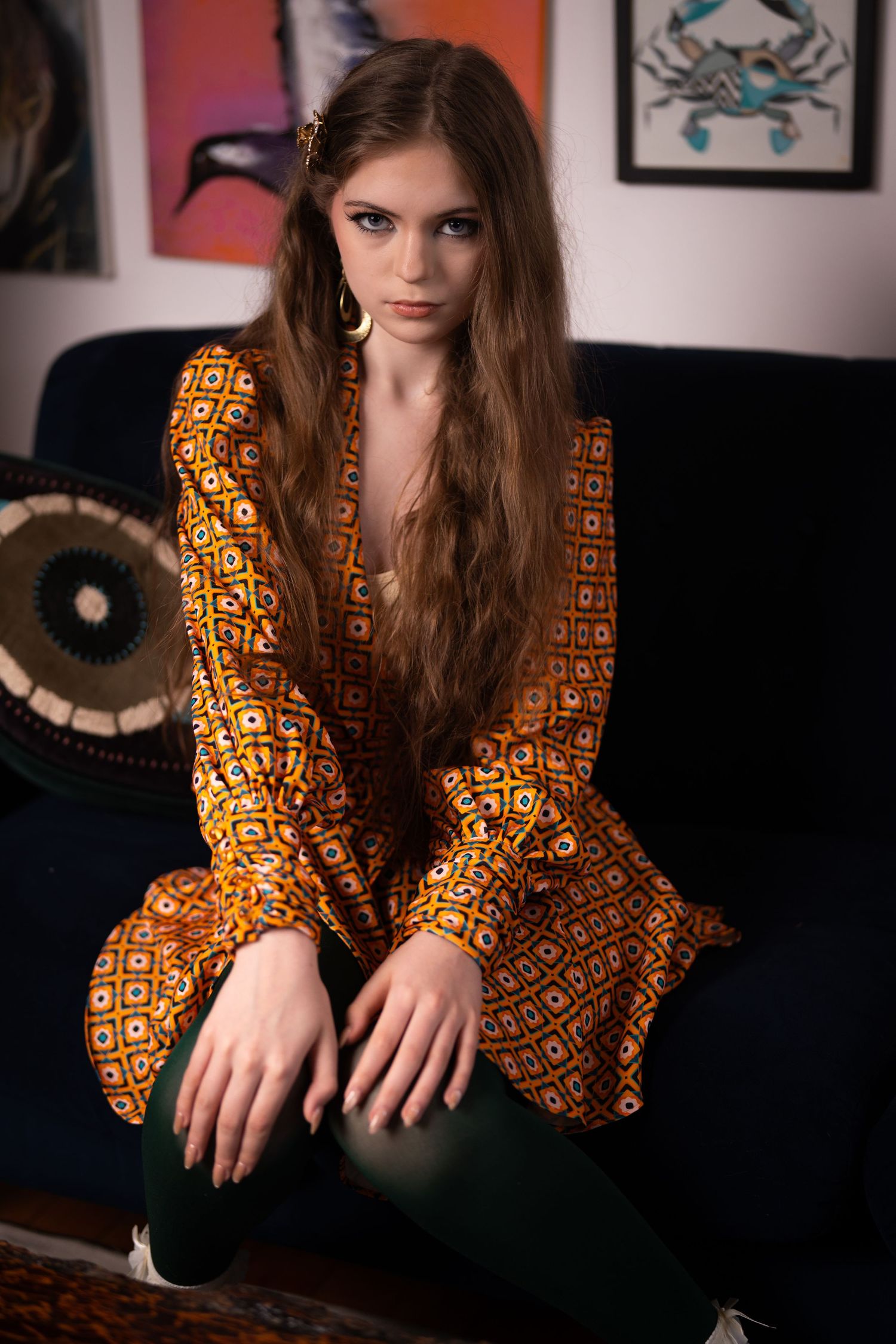 Young woman in bohemian orange patterned dress with green pants sits on dark sofa with eclectic art wall behind her.