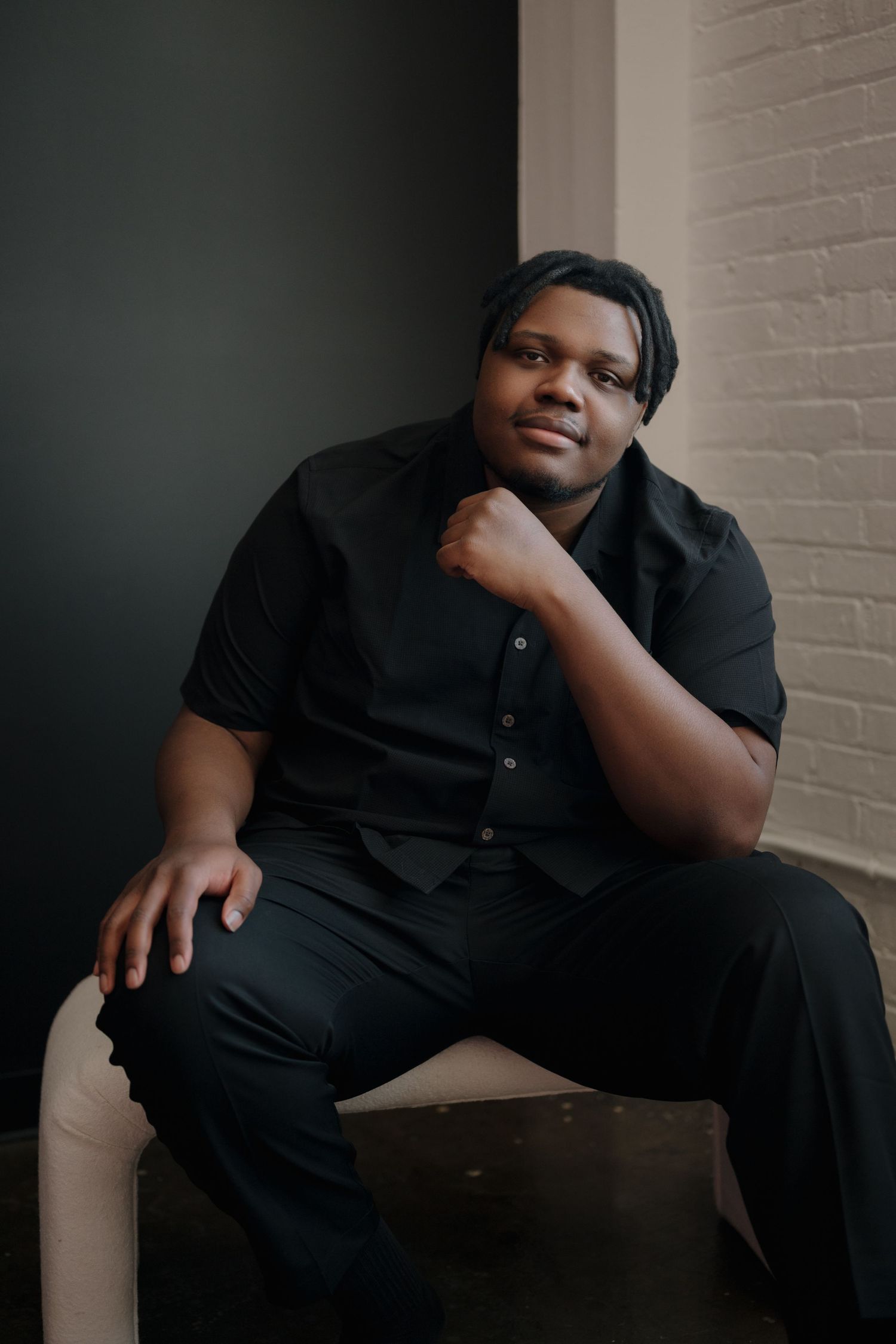 Young Black man in all-black outfit sitting confidently against a dark wall in a modern interior setting.