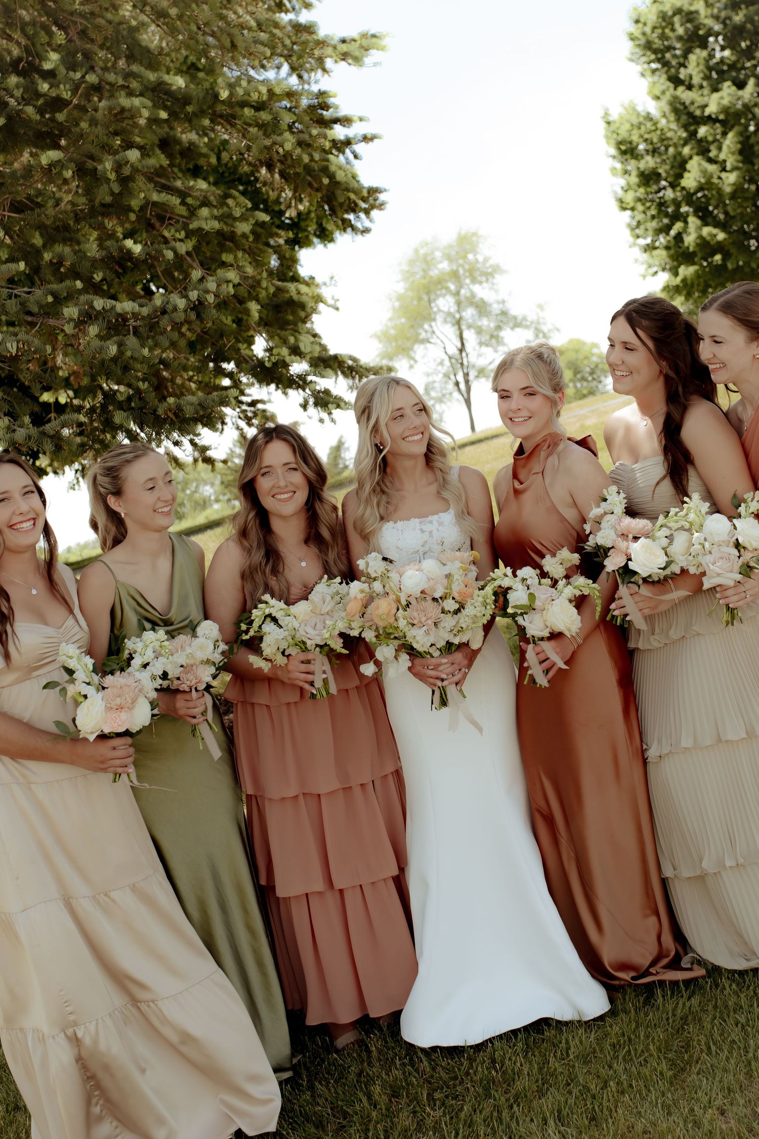 Bride in white gown poses with six bridesmaids in mismatched earth-tone dresses holding bouquets outdoors.