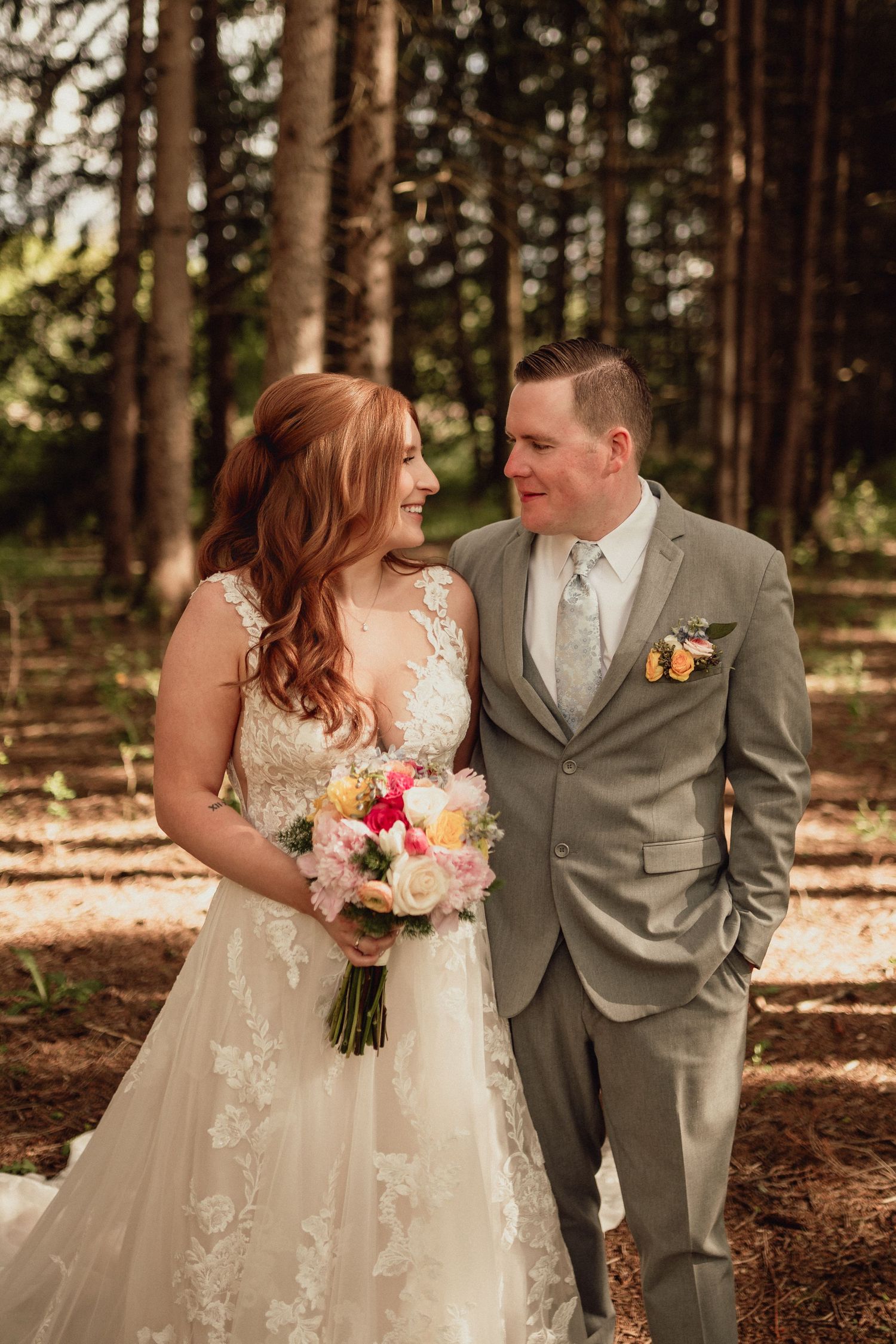 A couple in wedding attire shares intimate moments in a sunlit forest setting with pine trees in the background.