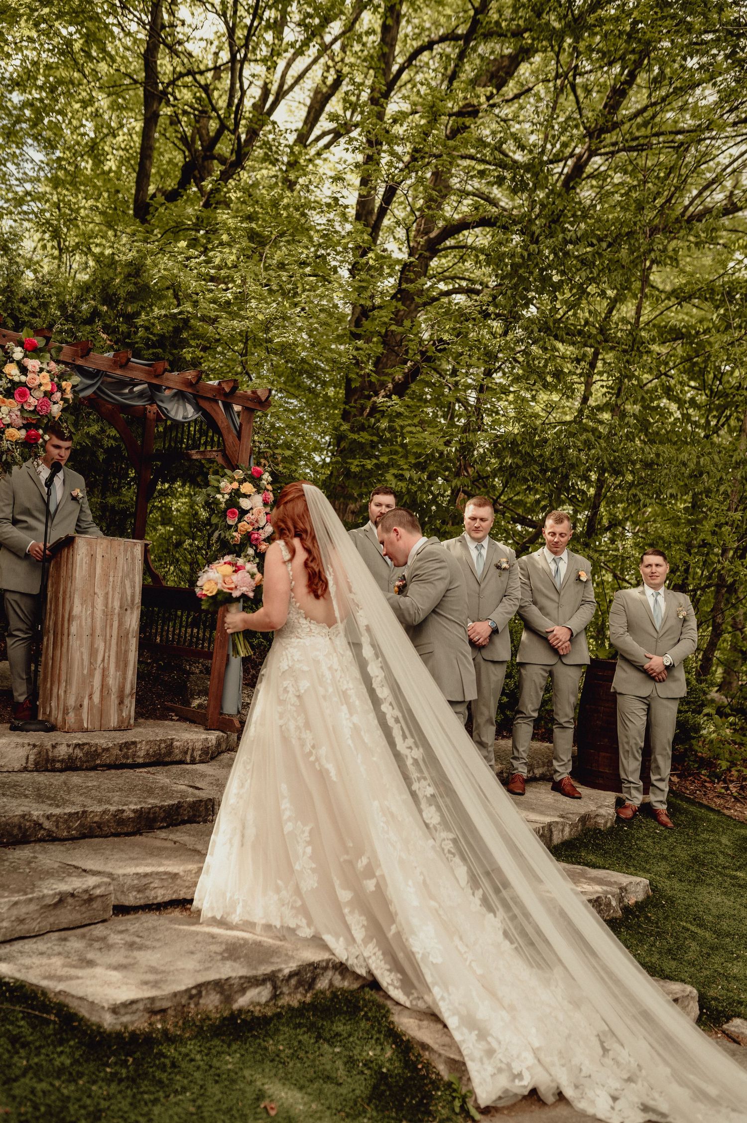 Wedding party poses in a garden setting with two brides wearing flowing white dresses and groomsmen in gray suits.