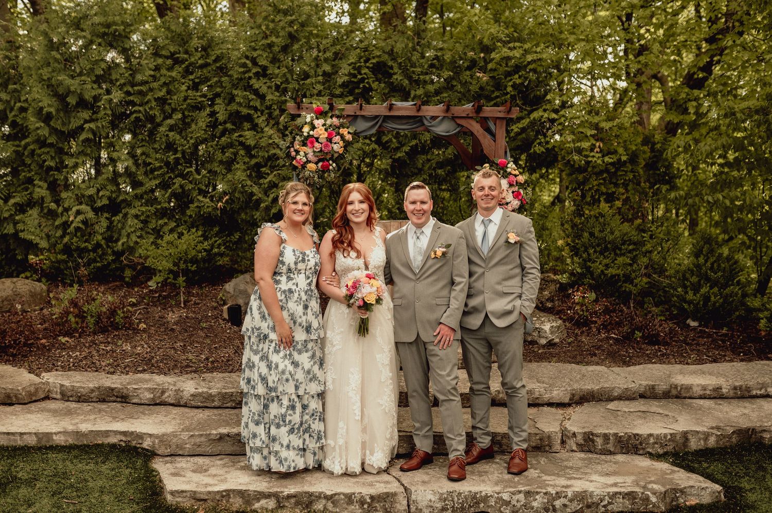 Wedding party in gray suits and floral dresses pose together in front of lush garden backdrop.