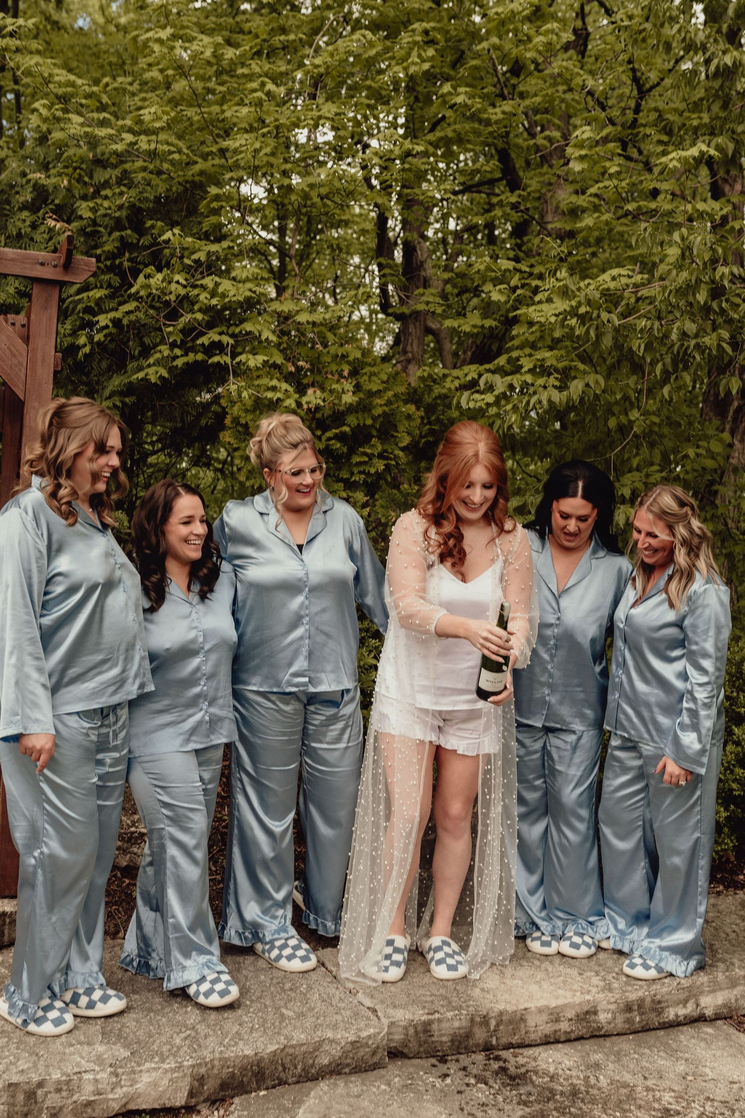 Wedding party wearing matching gray silk pajamas and fuzzy slippers poses together in a fun outdoor group photo.