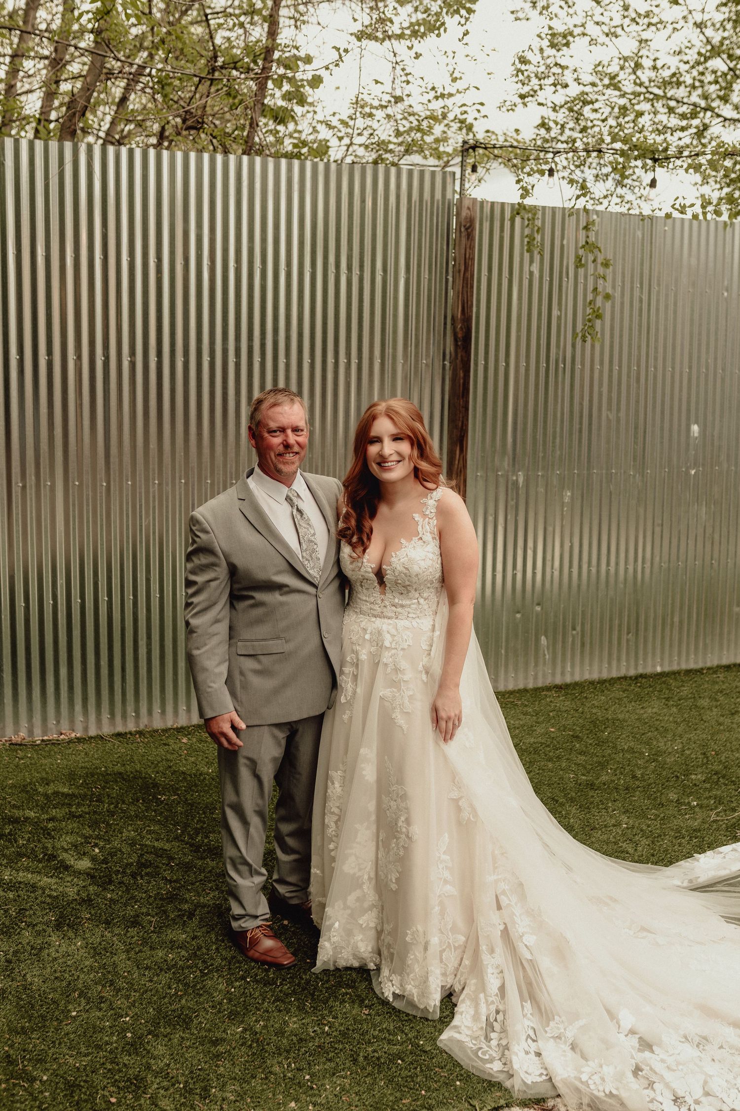 A wedding couple poses together against a metal wall backdrop in a garden setting with soft natural lighting.