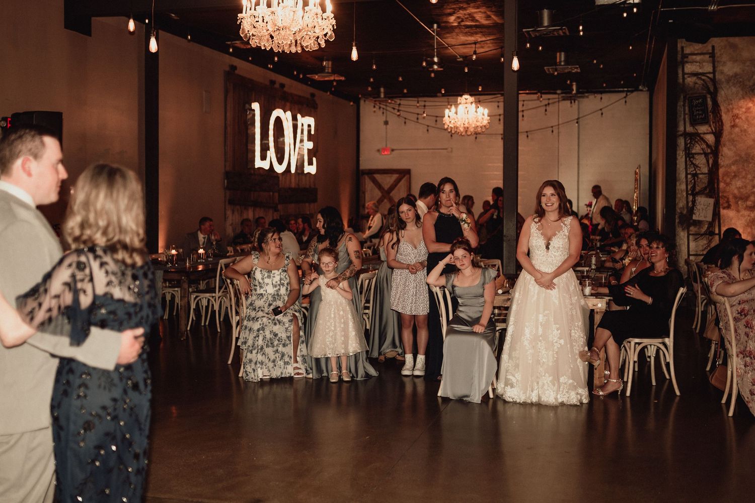 A wedding reception venue with a glowing LOVE sign, chandeliers, and guests dancing on the wooden floor.