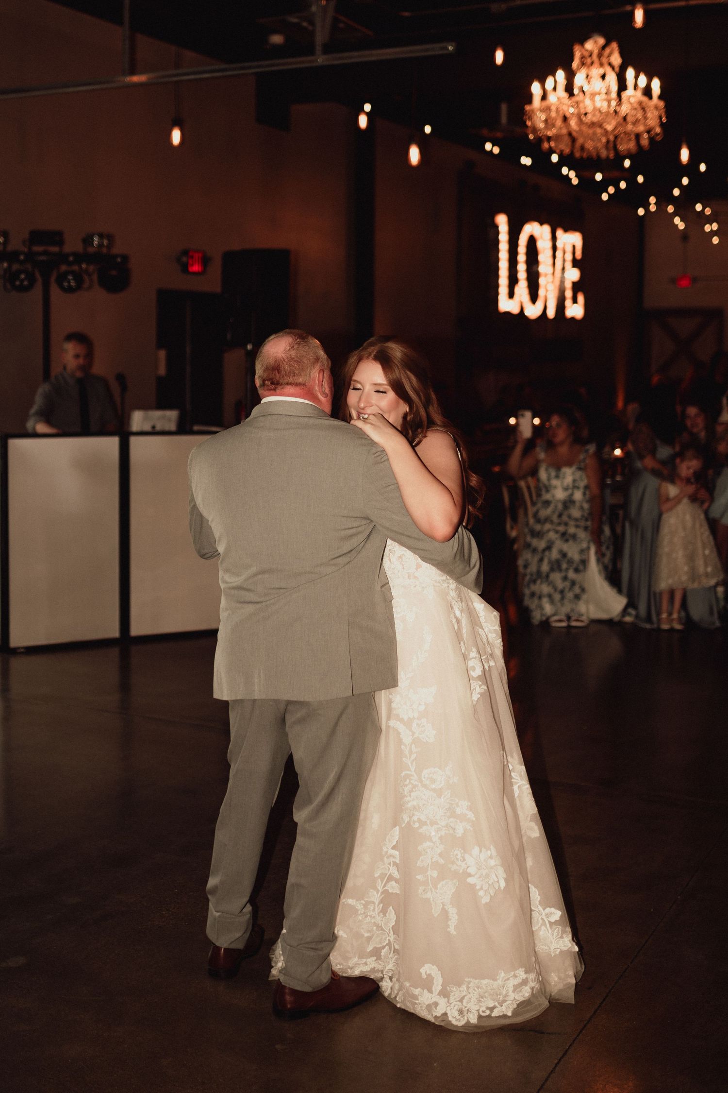 Newlywed couple dances together in a dimly lit reception venue with a chandelier and 'LOVE' sign glowing in the background.