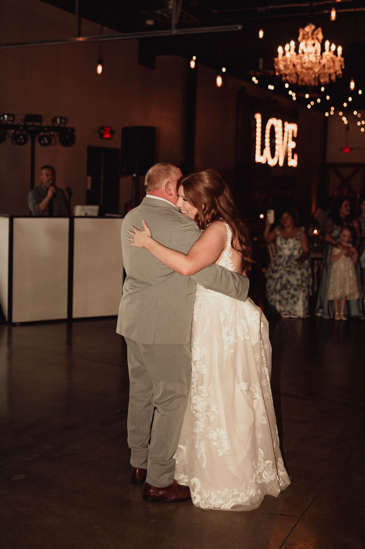 A couple shares their first dance at a wedding reception under a crystal chandelier in a dimly lit ballroom.