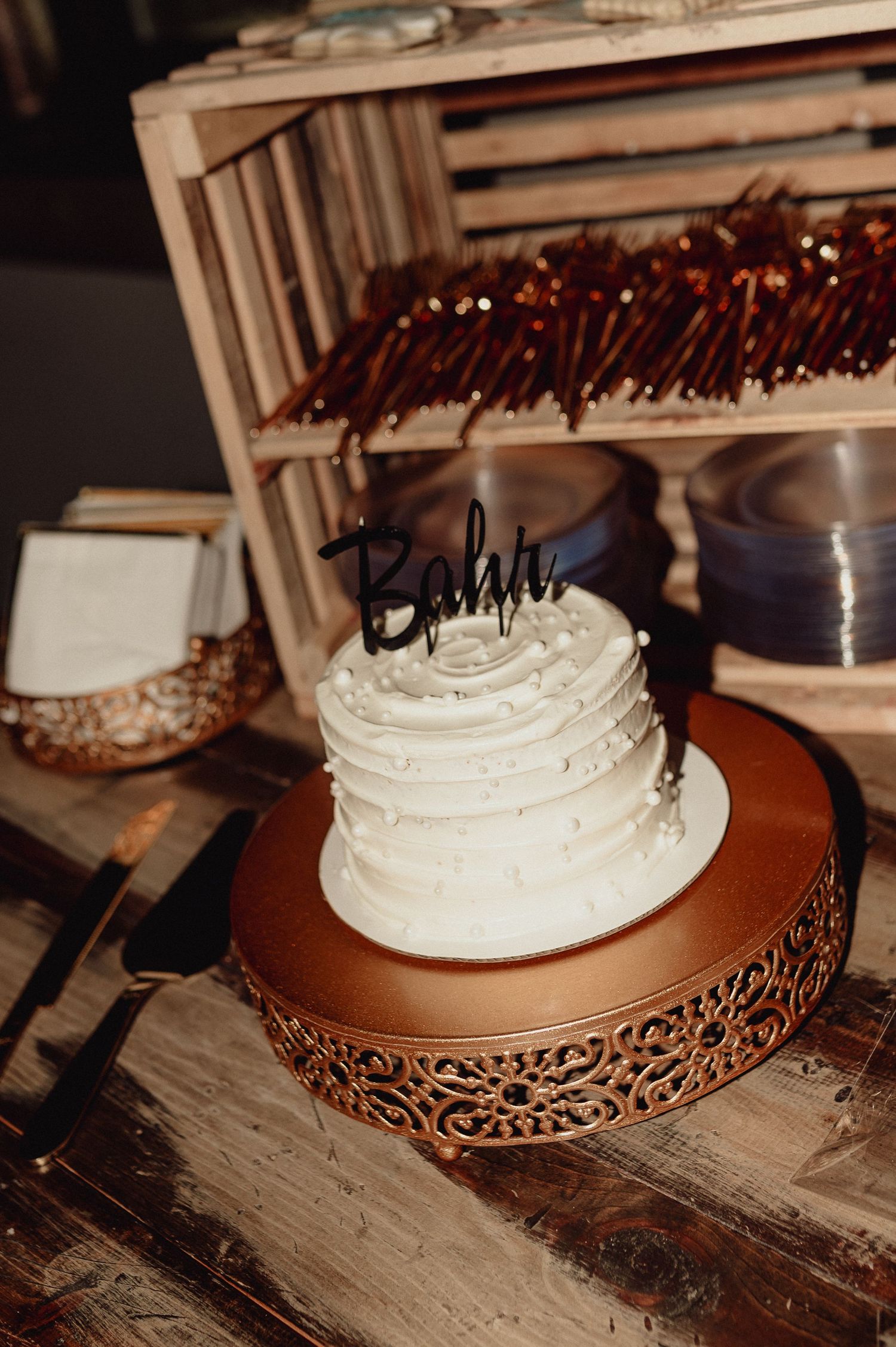A white frosted cake sits elegantly on a decorative copper-colored metal cake stand against a rustic wooden surface.