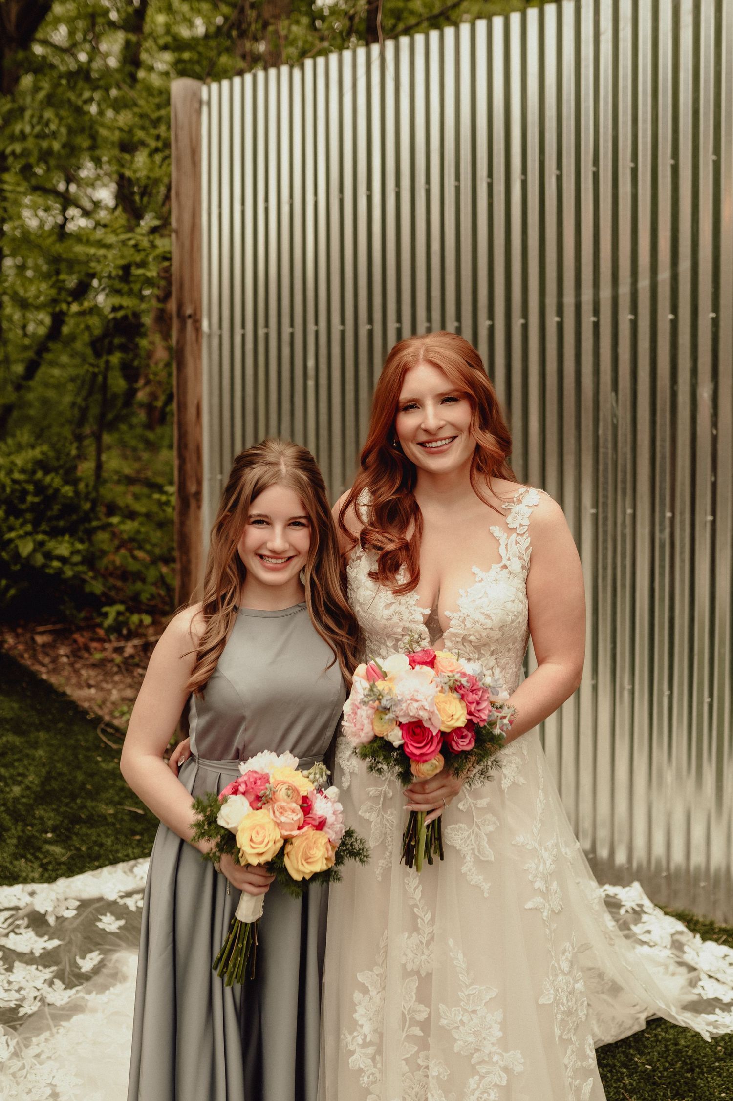 A wedding portrait showing a bridesmaid in gray and bride in lace gown holding colorful bouquets against metal wall.