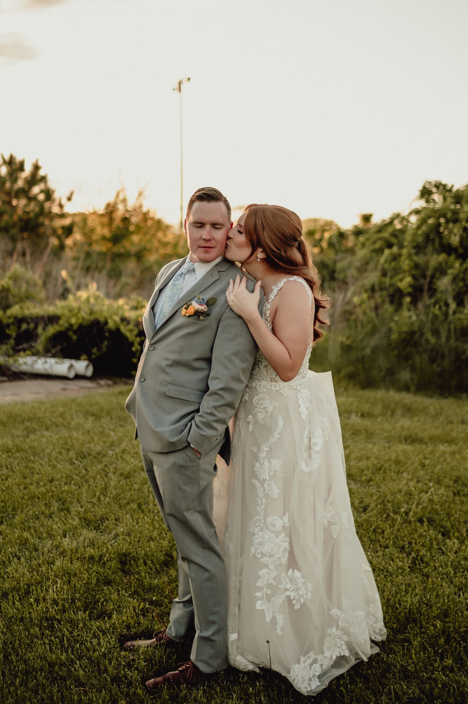 A couple in wedding attire shares an intimate moment outdoors at sunset, with the bride in white lace and groom in gray suit.