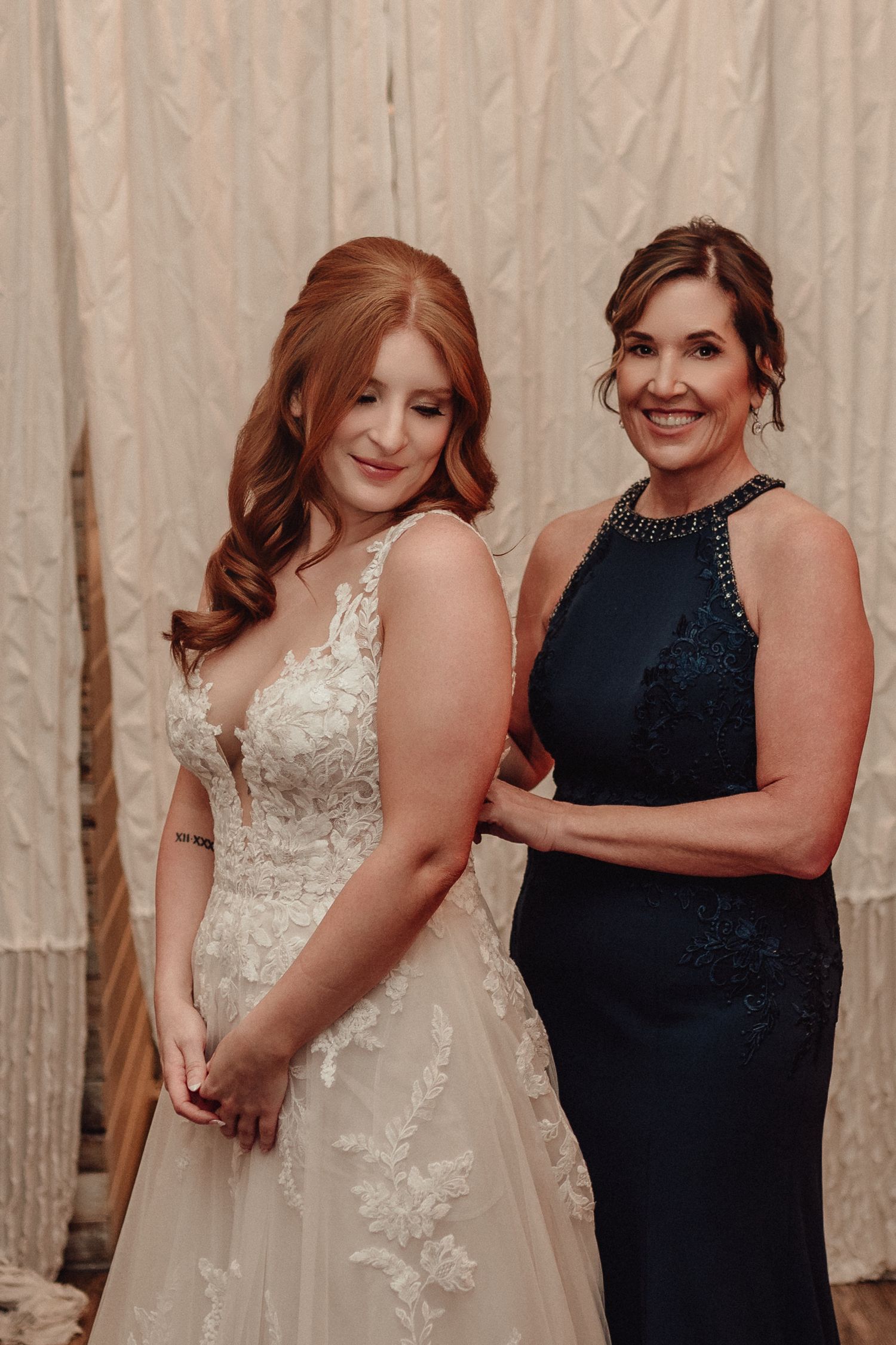 Two women pose together at a formal event, one wearing a white lace wedding gown and one in a navy blue dress.