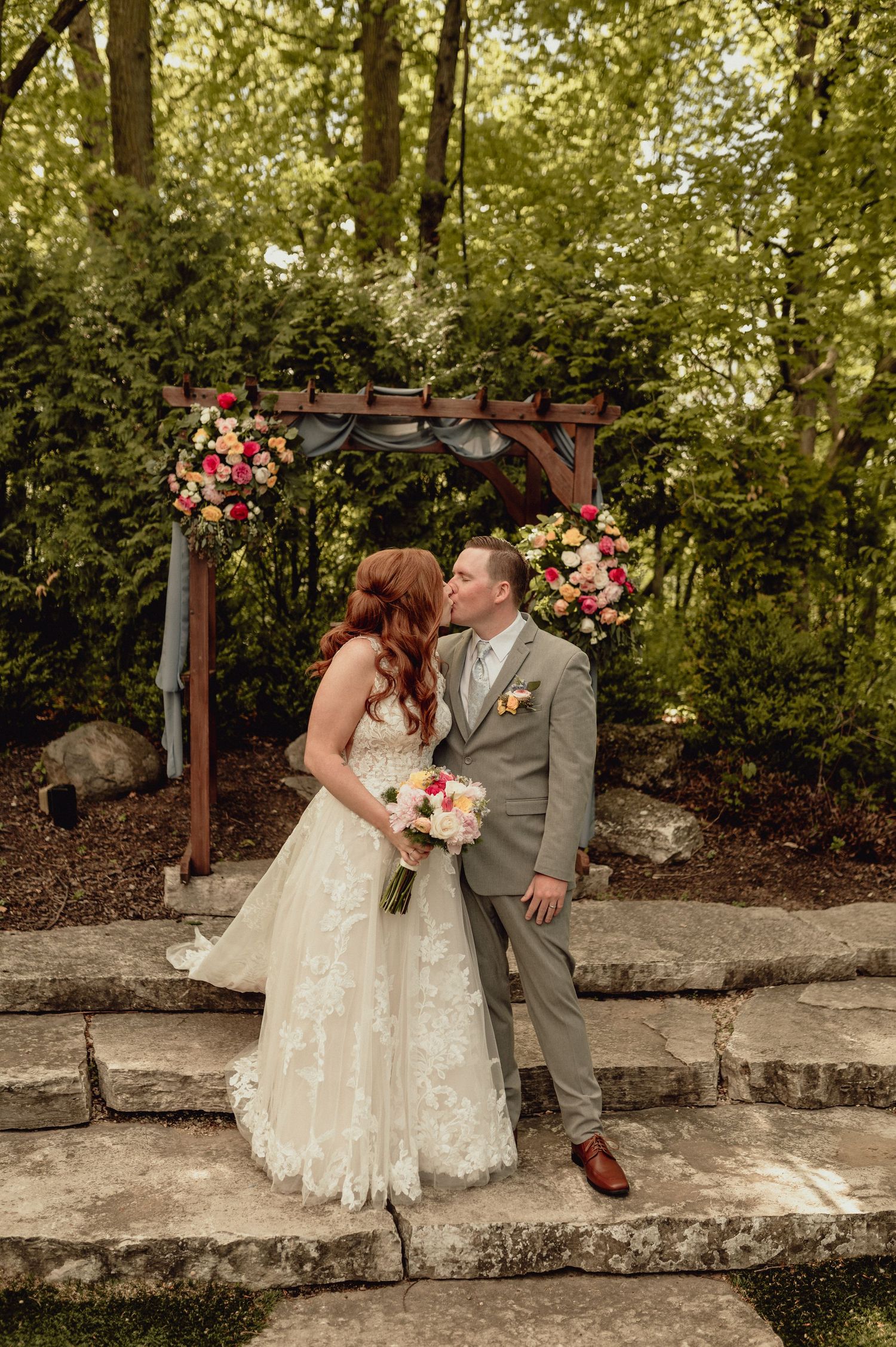 Romantic wedding couple shares tender moments on stone wall in garden setting with floral arch and greenery.