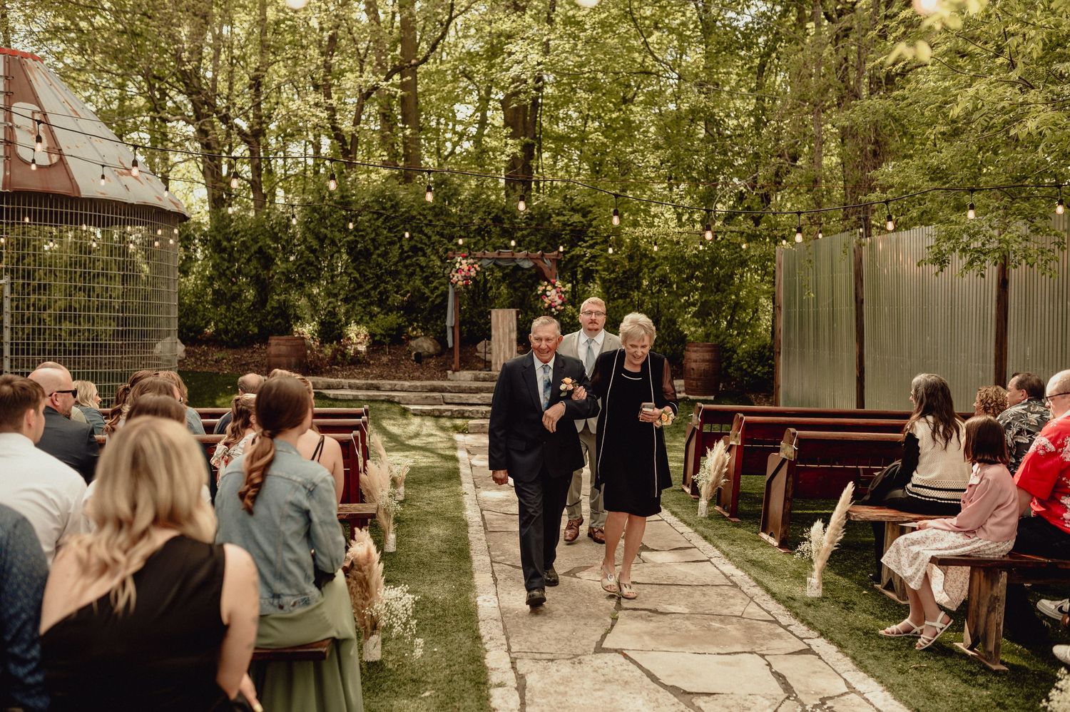 Wedding guests gather outdoors at a rustic venue with wooden benches and natural greenery during a ceremony.