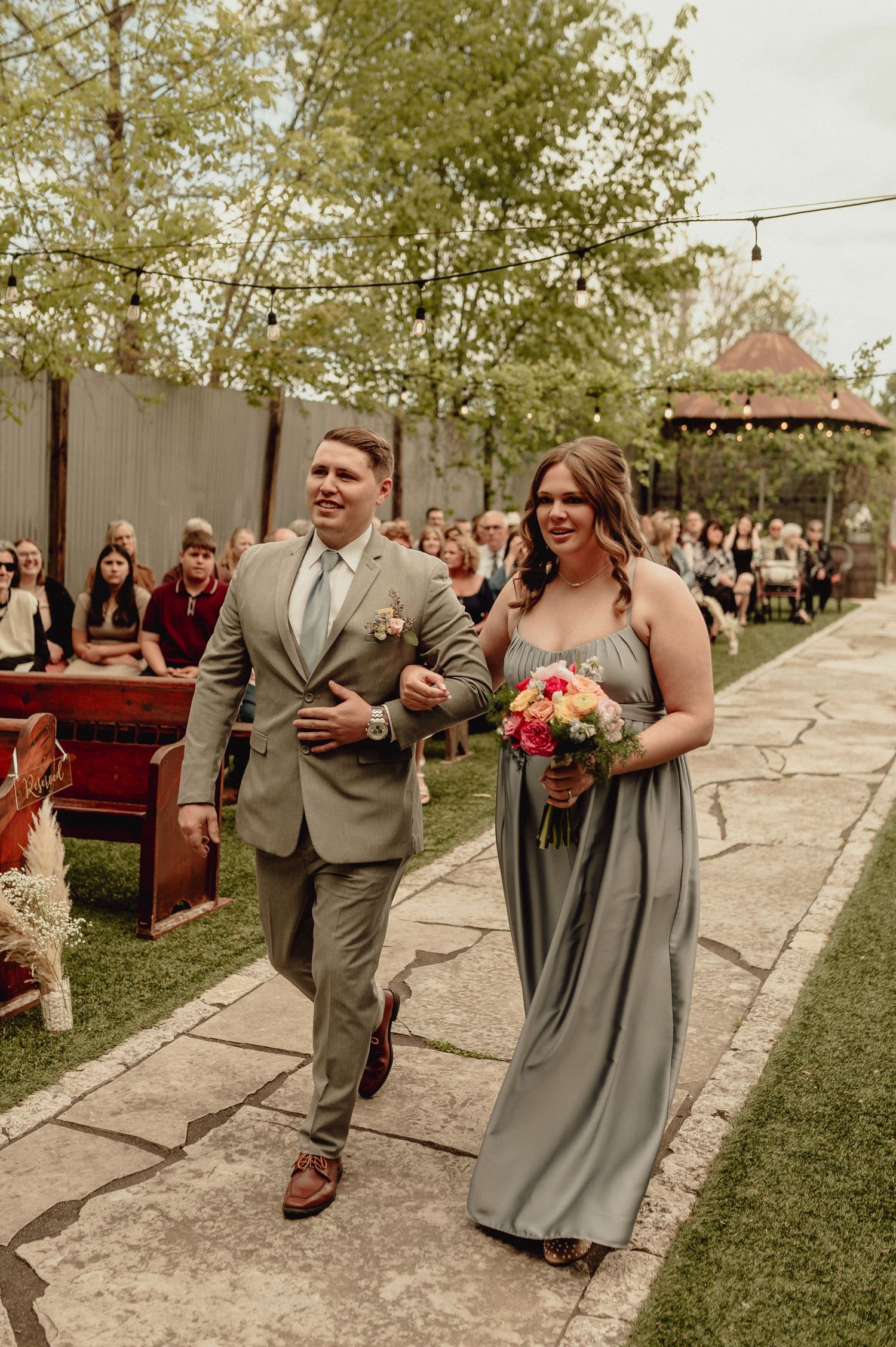 Wedding party members in gray dresses and tan suits walk and laugh together along an outdoor path at a celebration.