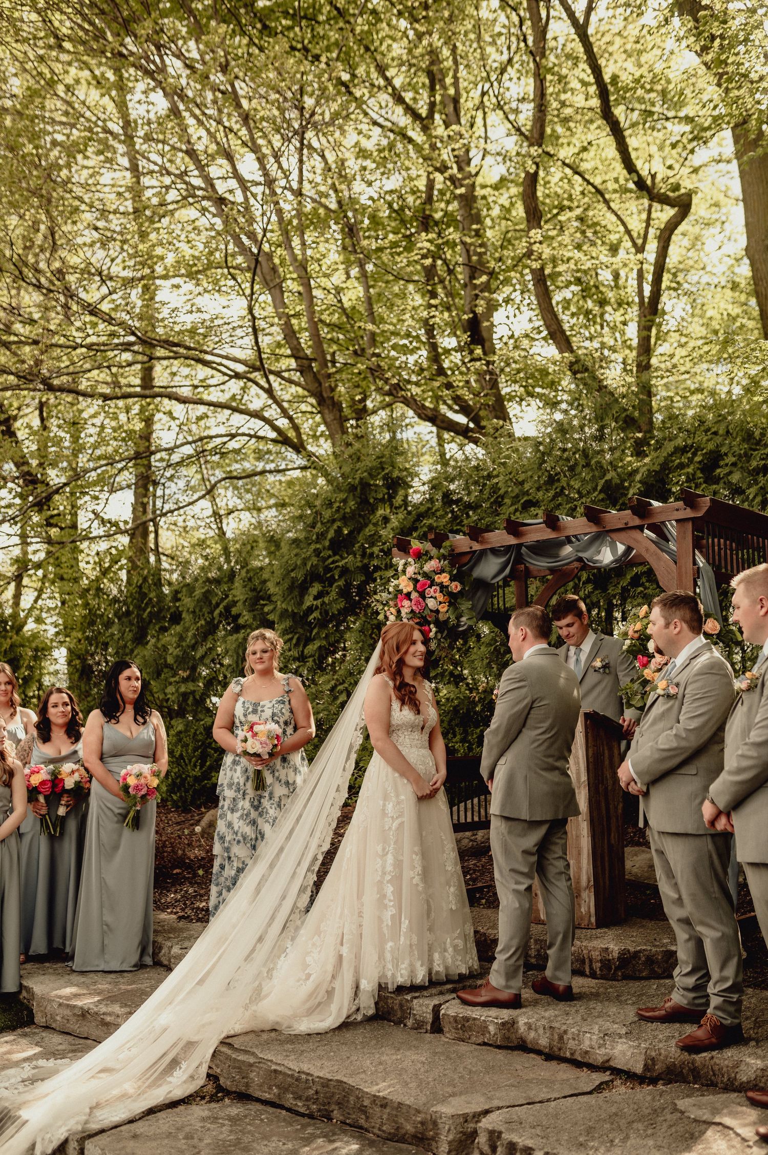 Wedding party in grey attire gathers under trees during outdoor ceremony with stone wall and floral decorations.