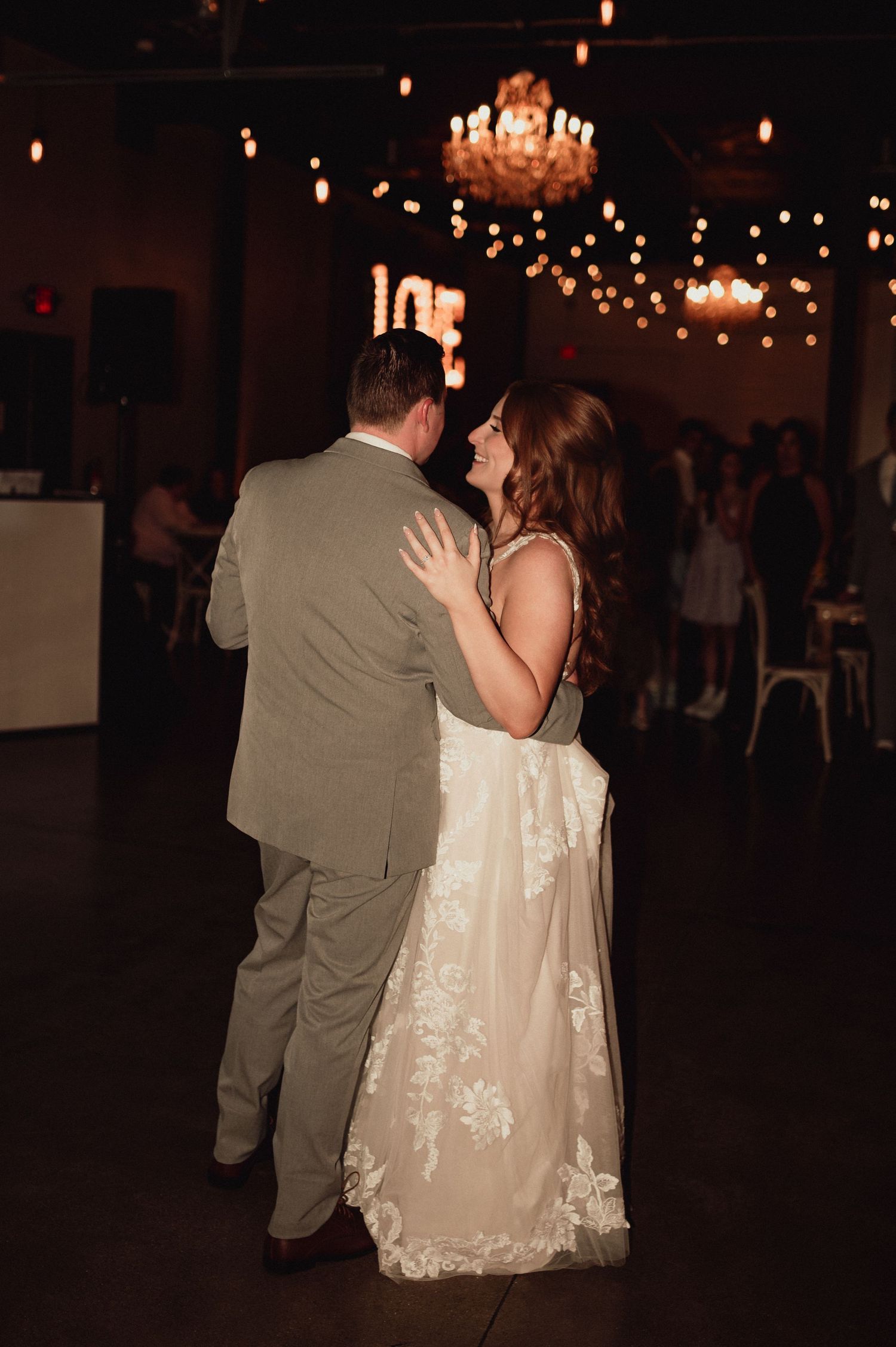 A couple shares a romantic dance moment at a dimly lit wedding reception with chandeliers overhead.
