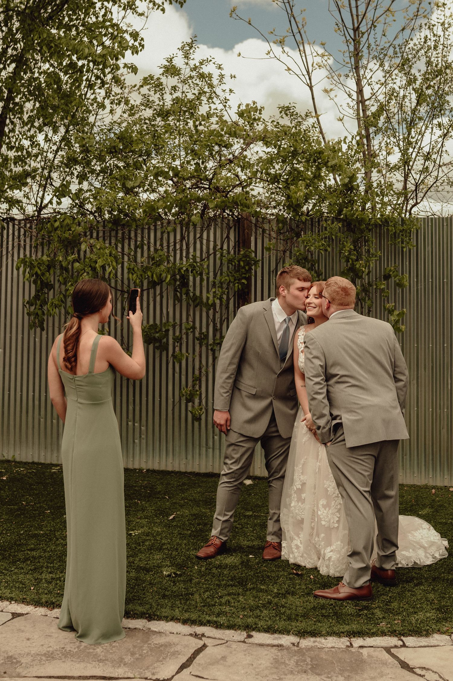 A wedding party gathers in front of a metal fence with greenery while a bridesmaid in sage green takes photos.