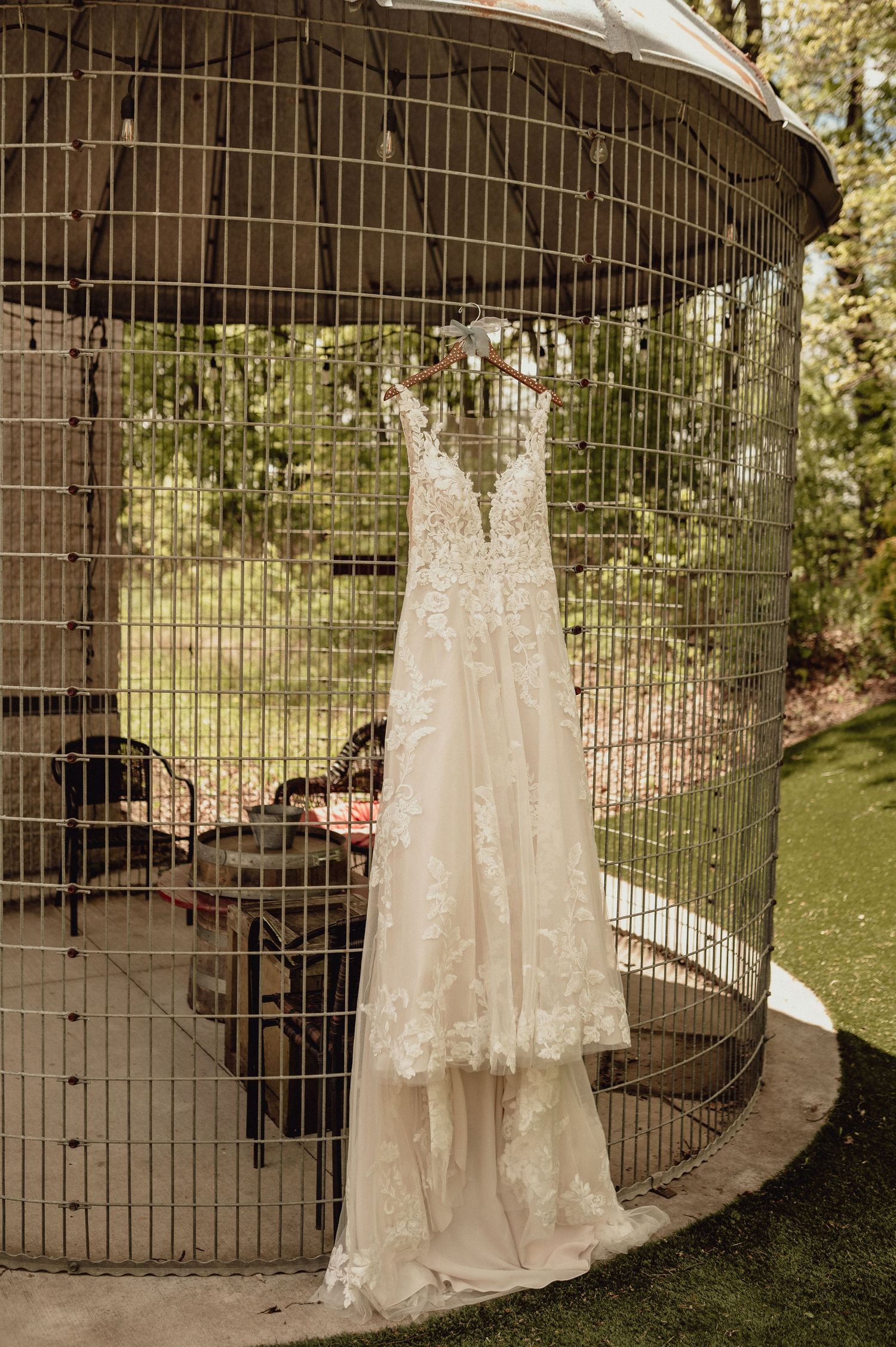 A delicate lace wedding dress hangs on a rustic metal gate in an outdoor garden setting.