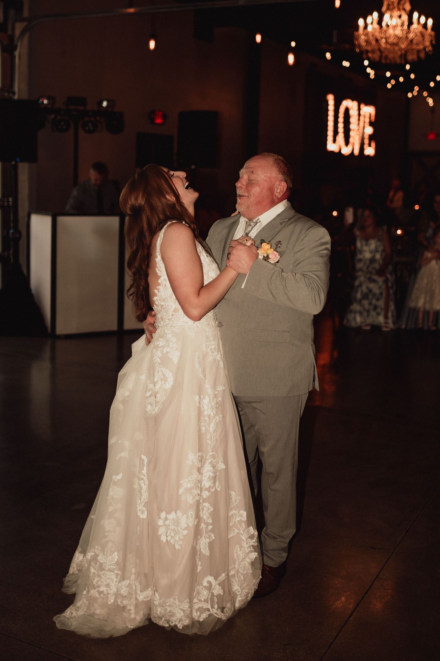 A couple shares a dance together at a wedding reception with chandeliers glowing overhead in a dimly lit ballroom.