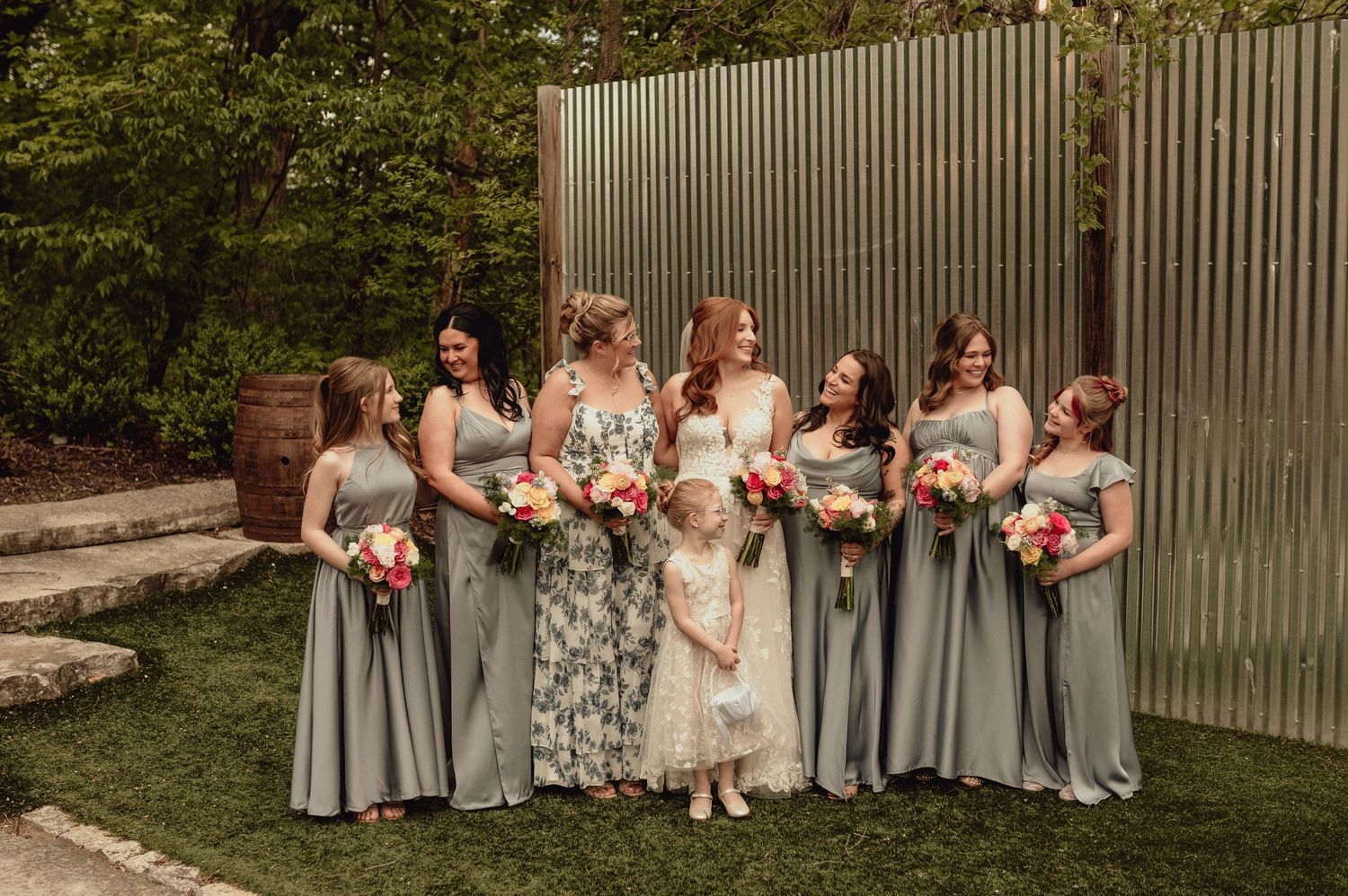 Wedding party in light gray dresses poses with coral and white bouquets in a garden setting.