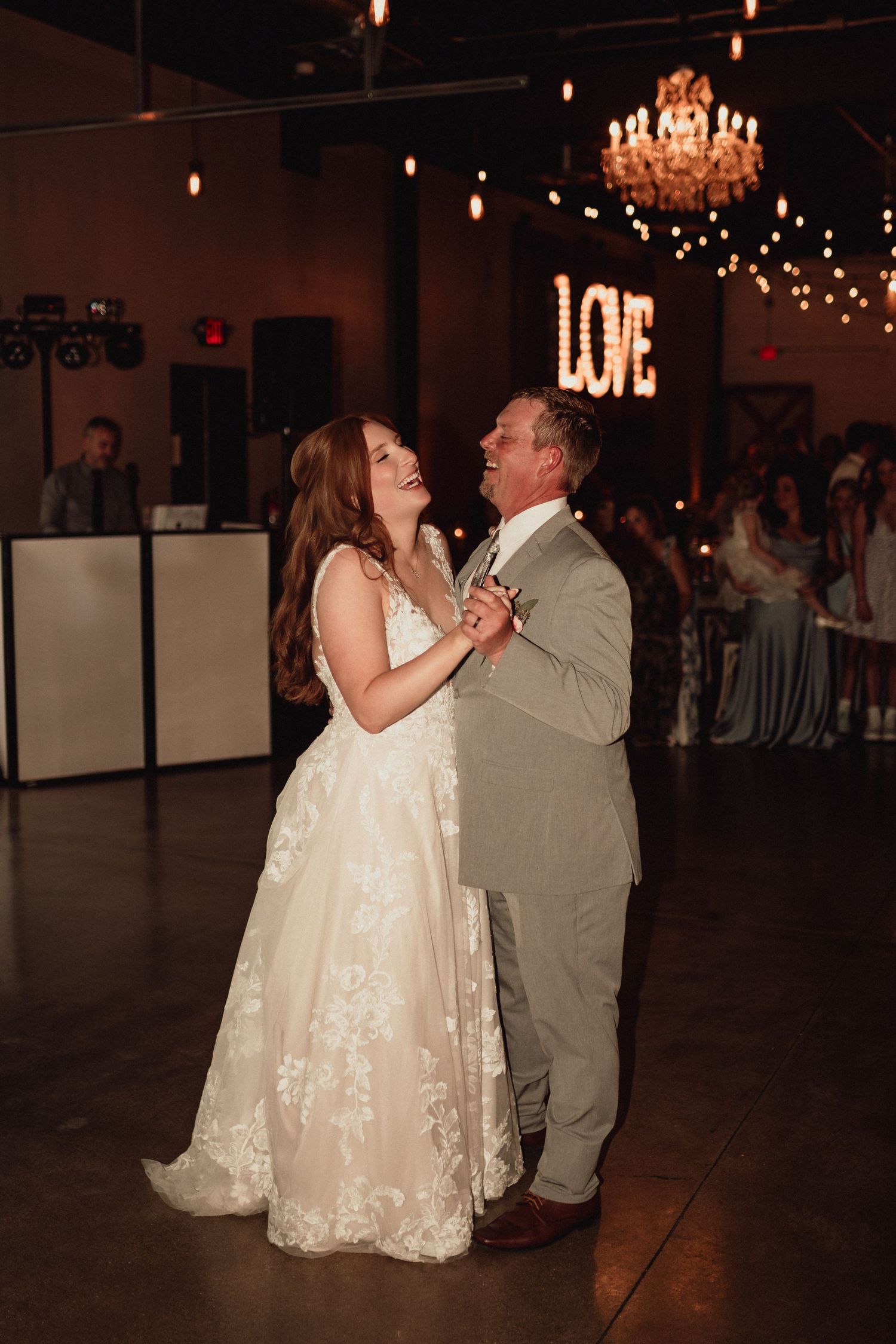 A couple shares their first dance as newlyweds under a chandelier in a dimly lit reception venue.