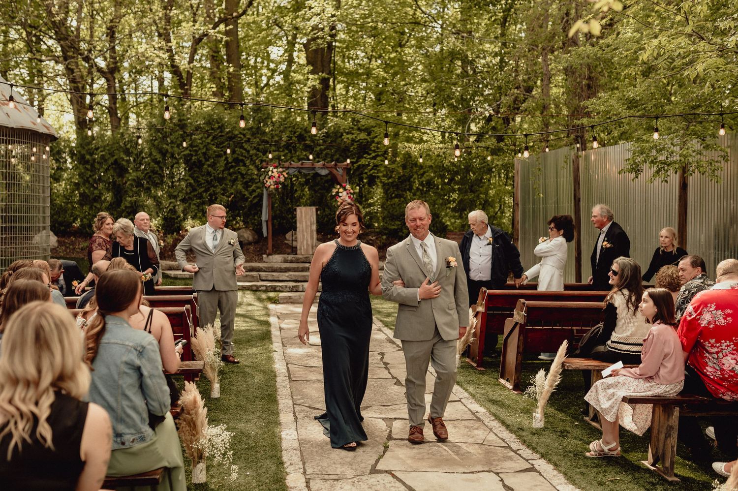 Wedding guests dance and celebrate on an outdoor patio surrounded by trees and string lights during a reception.