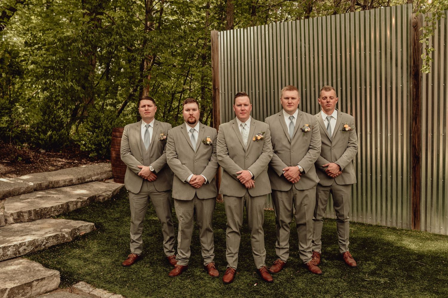 A wedding party in gray suits sits casually on stone steps against a corrugated metal wall.