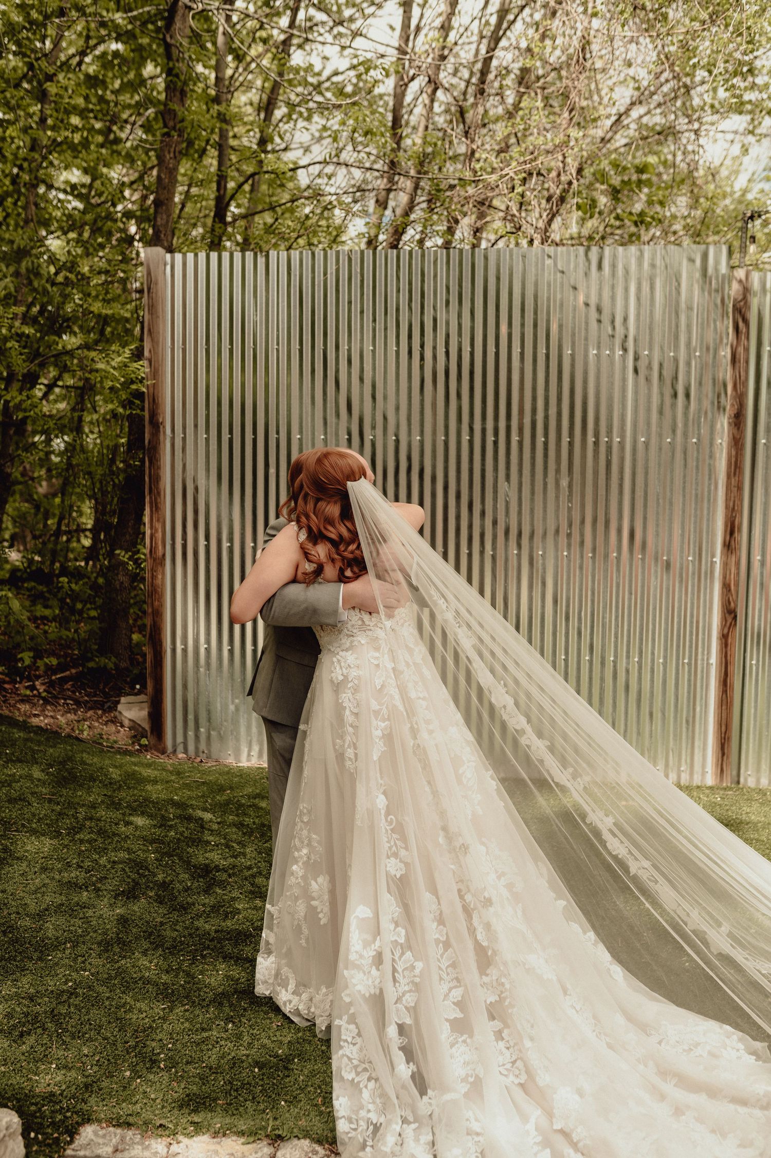 A wedding dress with a flowing lace train and cathedral veil is photographed against a rustic wooden fence backdrop.