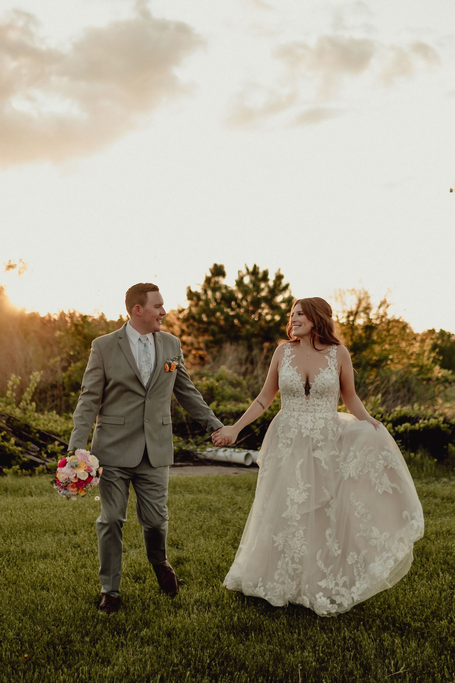 Bride and groom walk together at sunset in a grassy field while holding a bouquet of pink flowers.