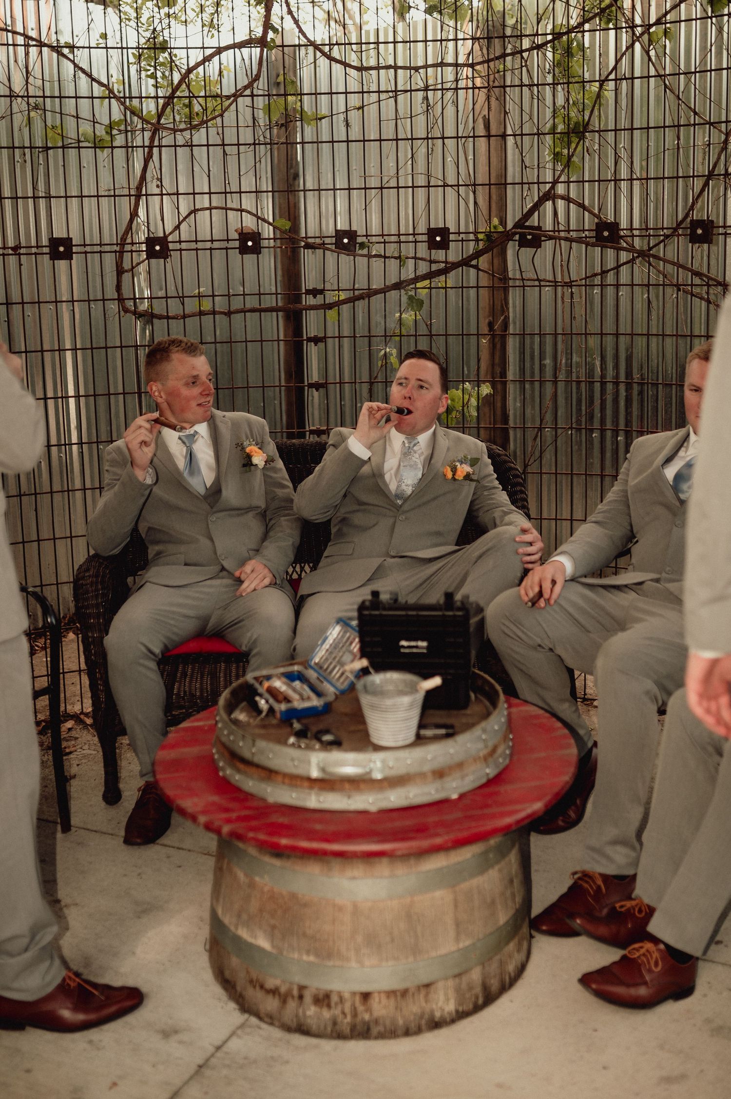 A group of groomsmen in gray suits sit on rustic wooden barrels during a wedding photoshoot.