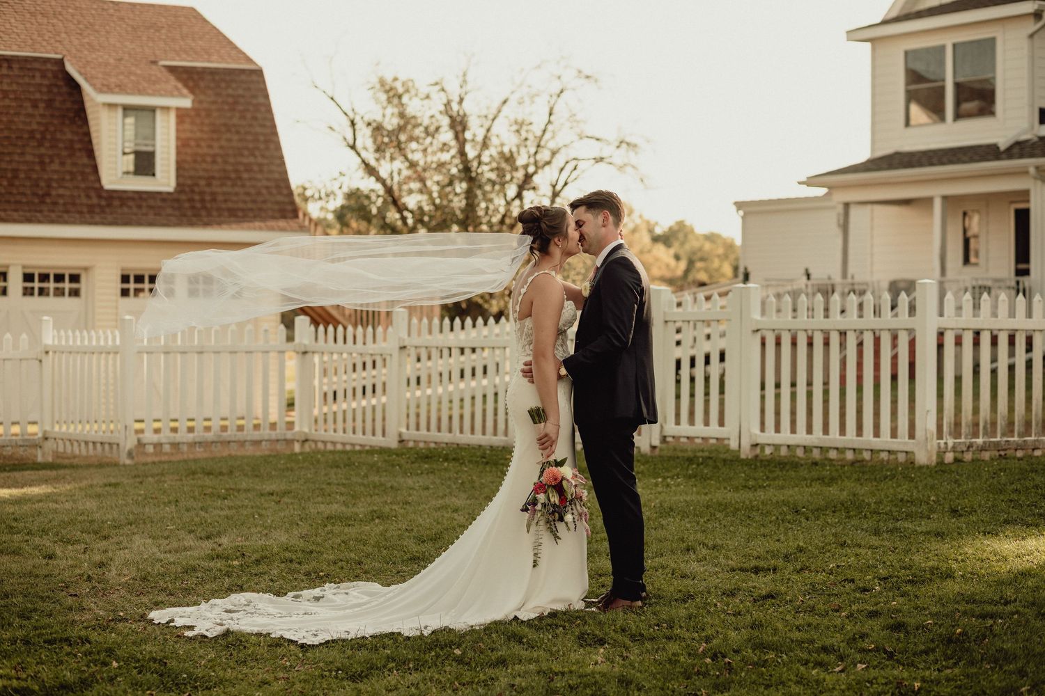 Wedding couple poses in front of white houses with picket fences at sunset.