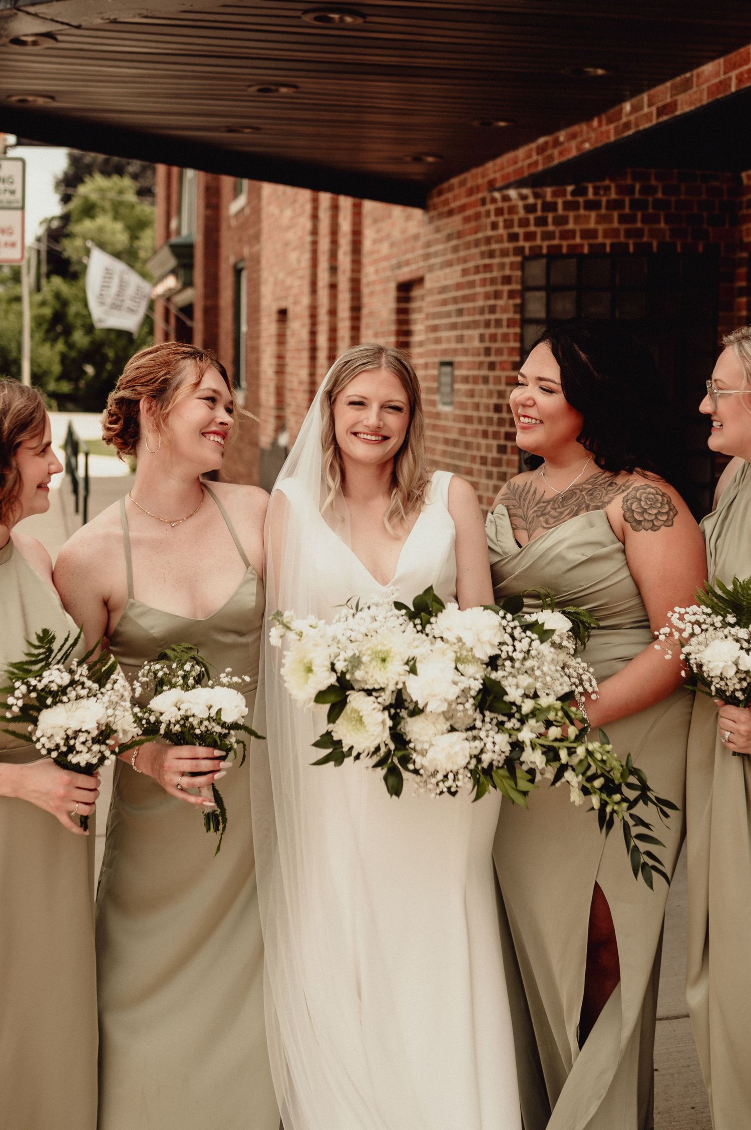 Bridesmaids in sage green dresses hold white bouquets while sharing laughs outside a brick church.
