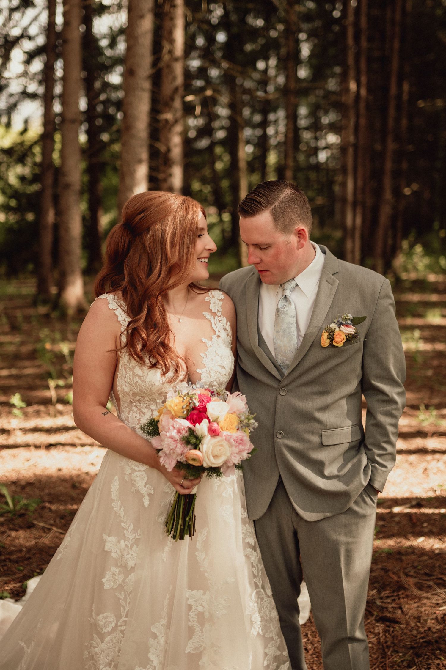 A couple in wedding attire share an intimate moment in a forest setting with sunlight filtering through pine trees.