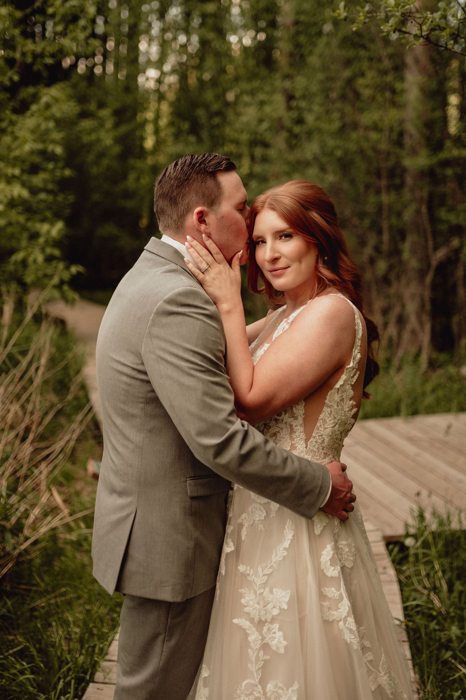 A romantic wedding couple shares intimate moments in a wooded outdoor setting wearing grey suit and lace gown.