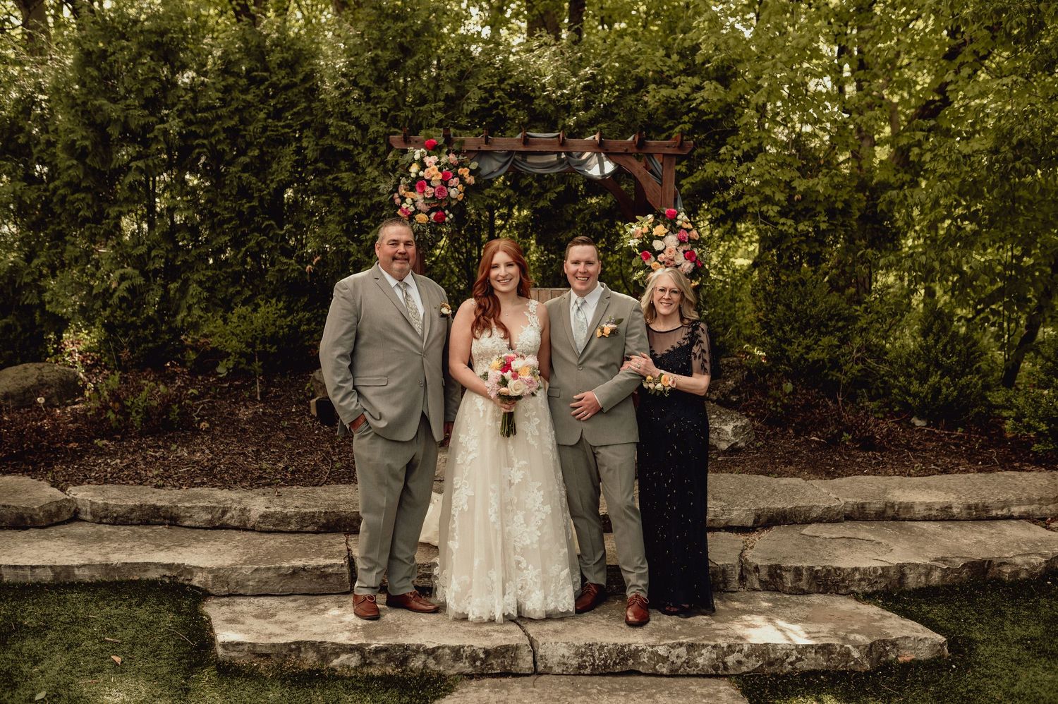 Wedding party poses together on stone wall in outdoor garden setting during summer celebration.