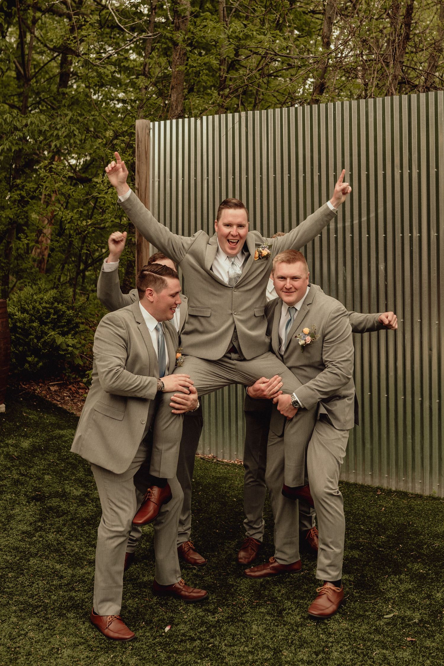 Four playful groomsmen in gray suits strike fun poses against a fence in an outdoor garden setting.