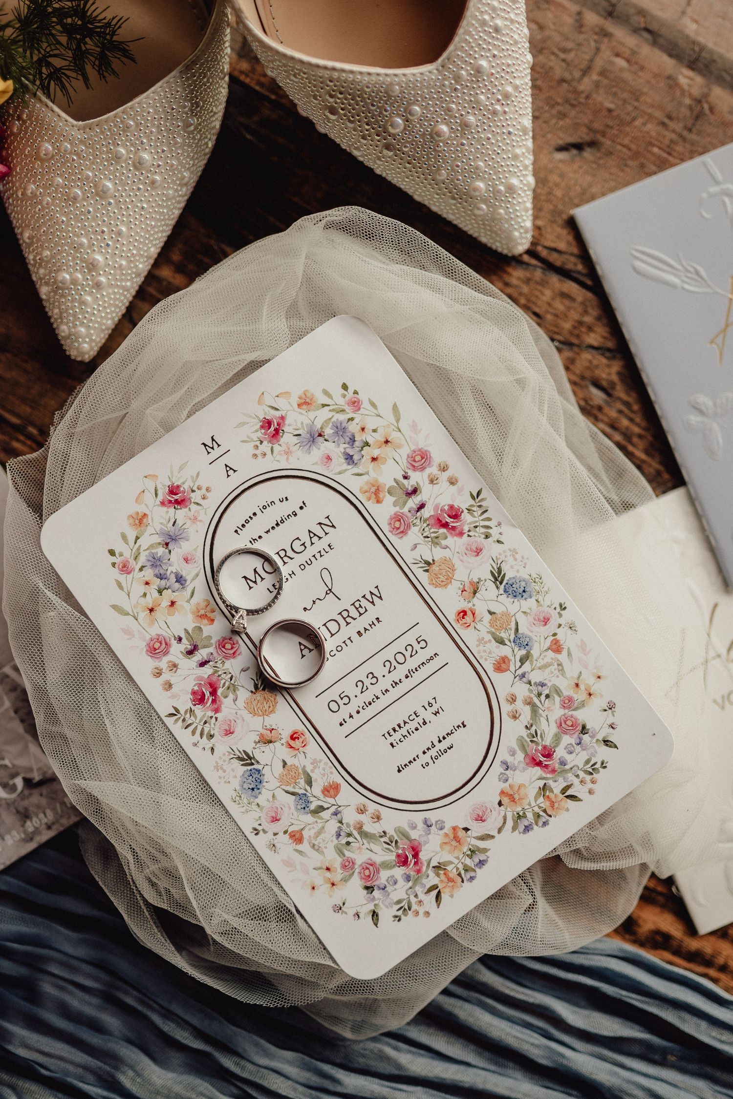 Wedding rings rest on a floral invitation card with delicate tulle fabric and beige bridal shoes on wooden surface.