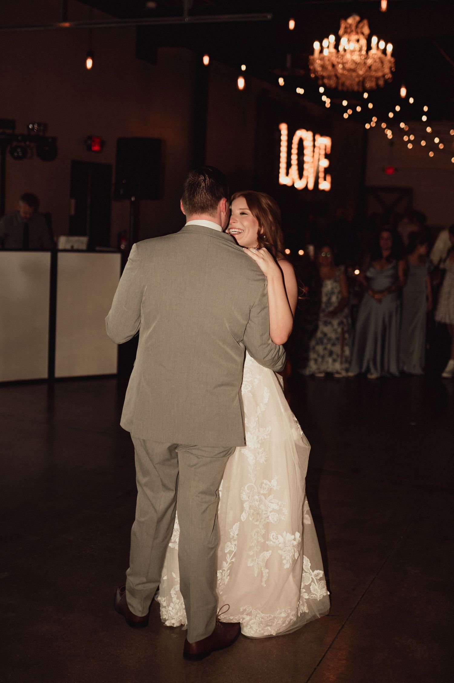 A couple dances together at a wedding reception under a crystal chandelier and twinkling lights.