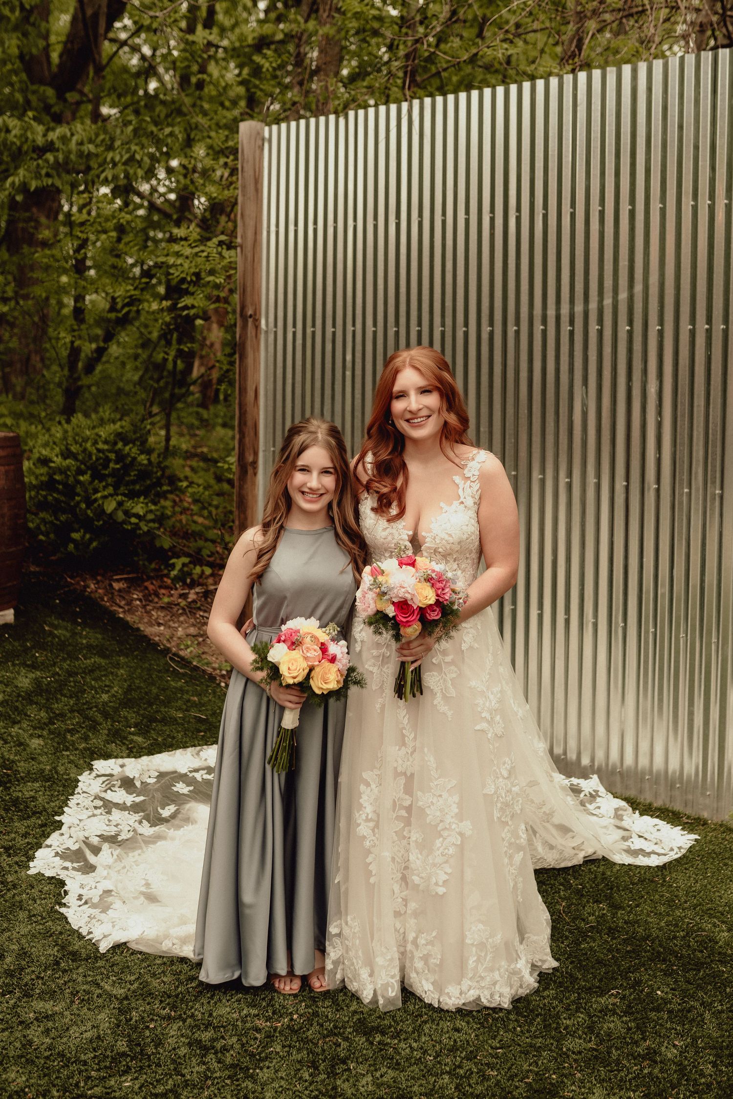 Two women pose outdoors in formal attire holding floral bouquets against a corrugated metal backdrop.