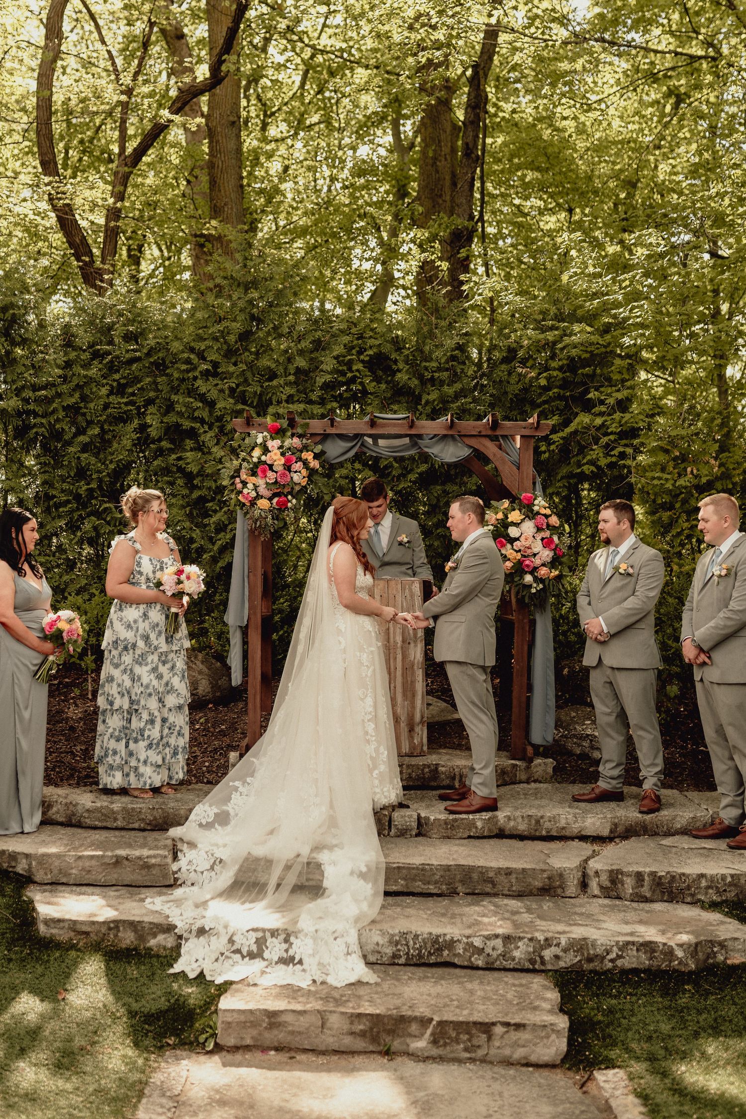 Wedding party stands in a line outdoors under trees with a rustic wooden pergola decorated with pink flowers.