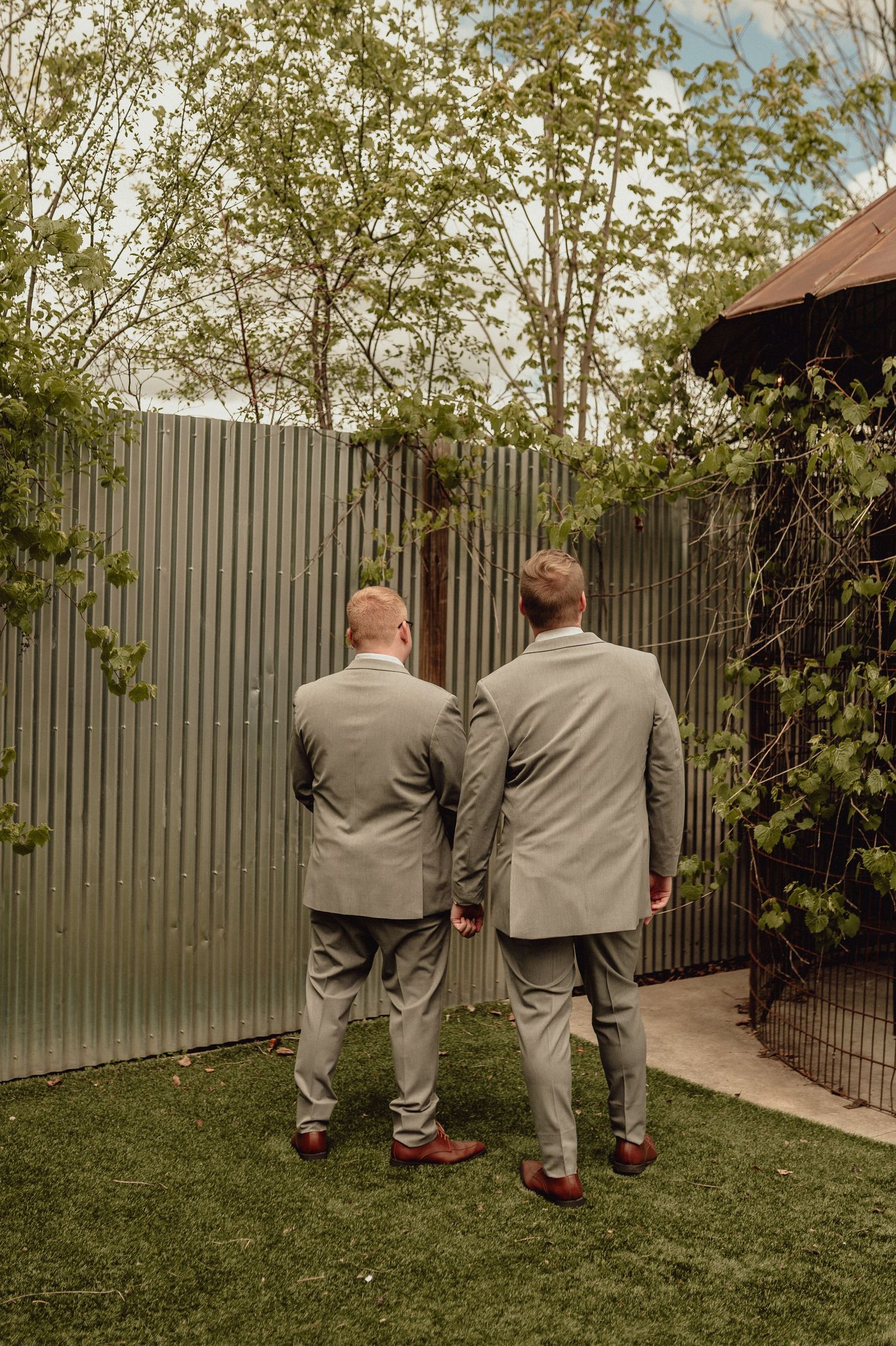 Two people in matching gray suits stand together in a backyard garden with a metal fence and trees in the background.