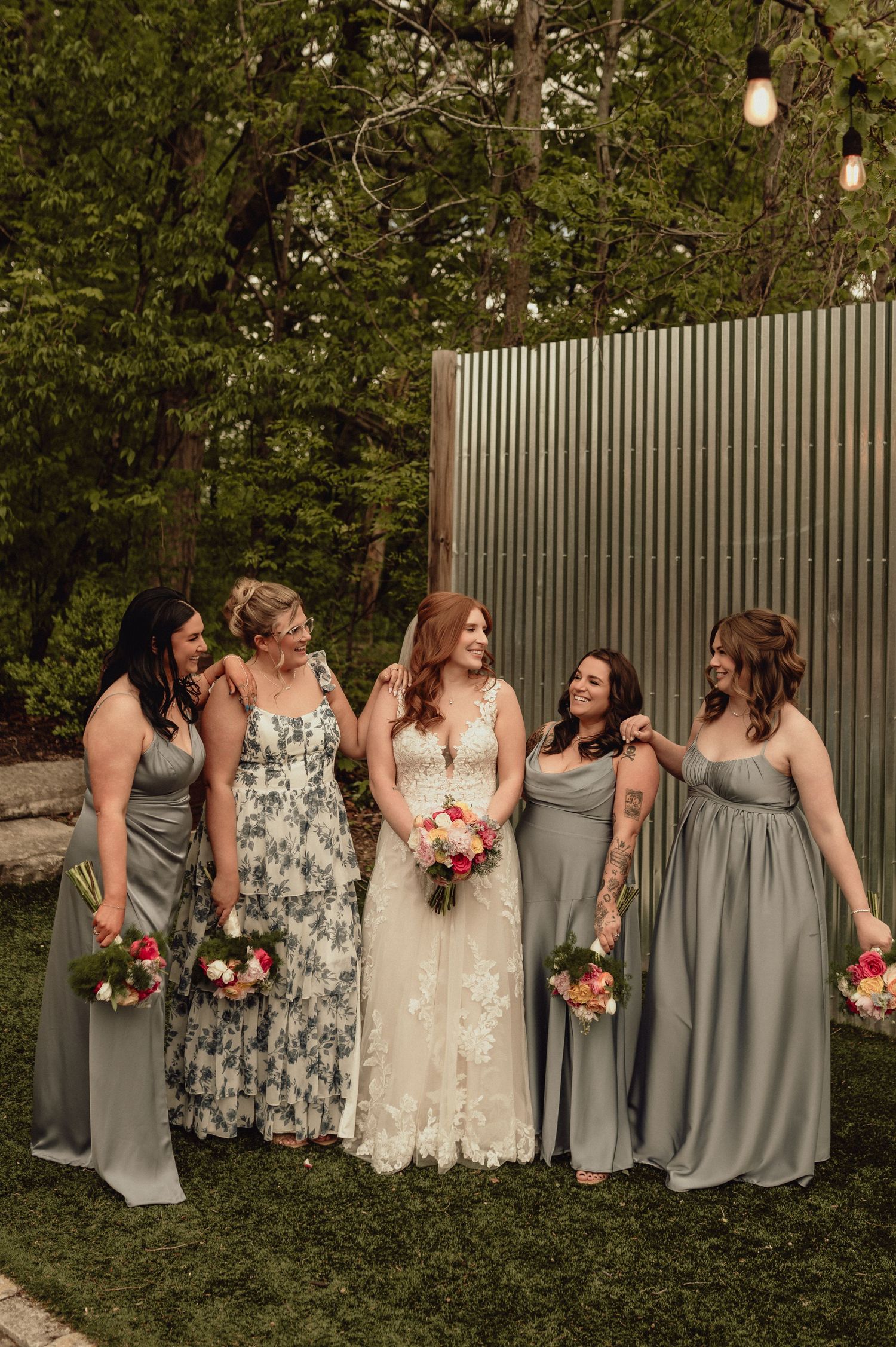 Bridal party in gray dresses and floral prints pose together in a garden setting with string lights overhead.