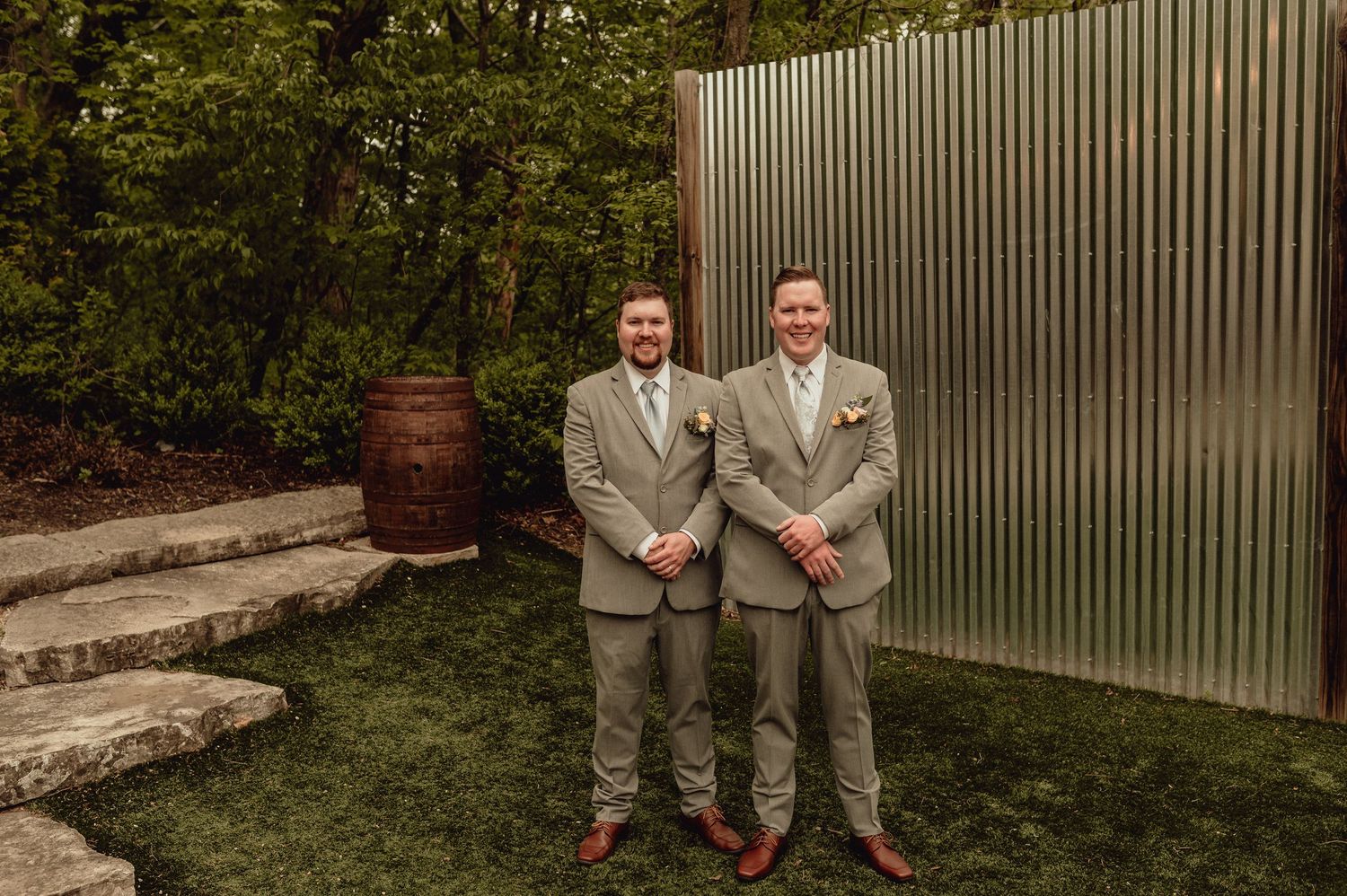 Two people in matching gray suits stand together in a garden setting with a wooden fence and greenery in the background.